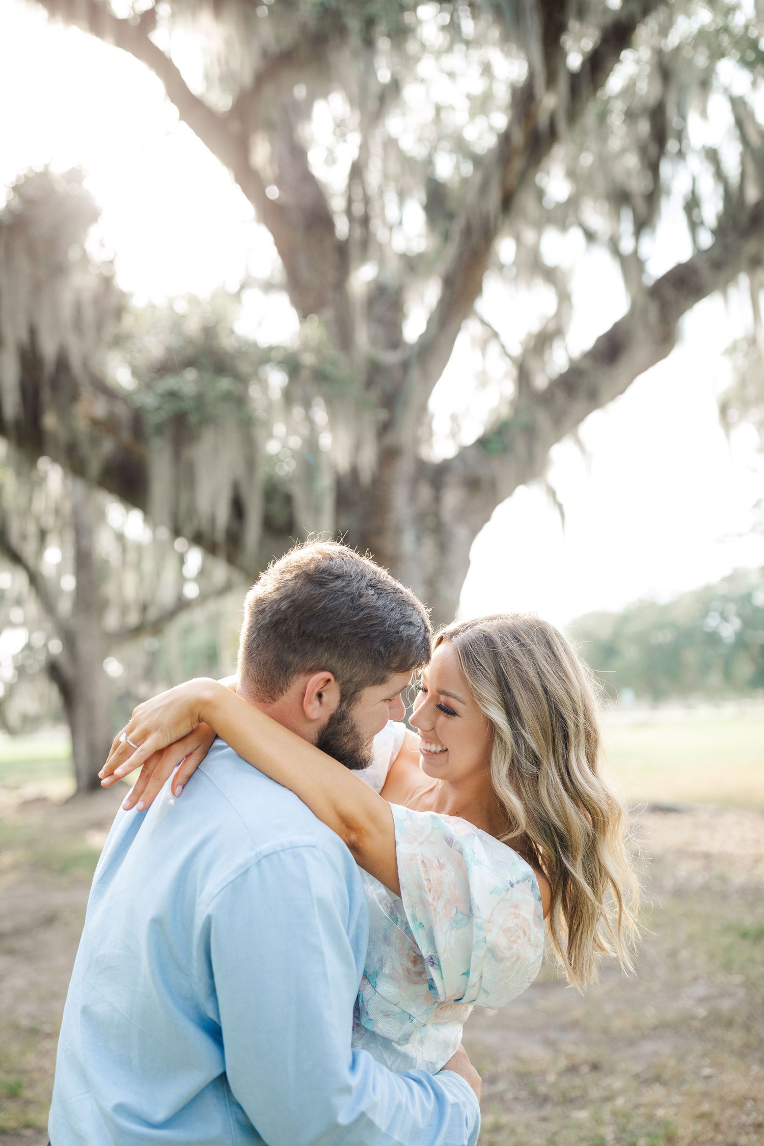 A couple embraces and smiles at each other beneath Spanish moss hanging from majestic oak trees.