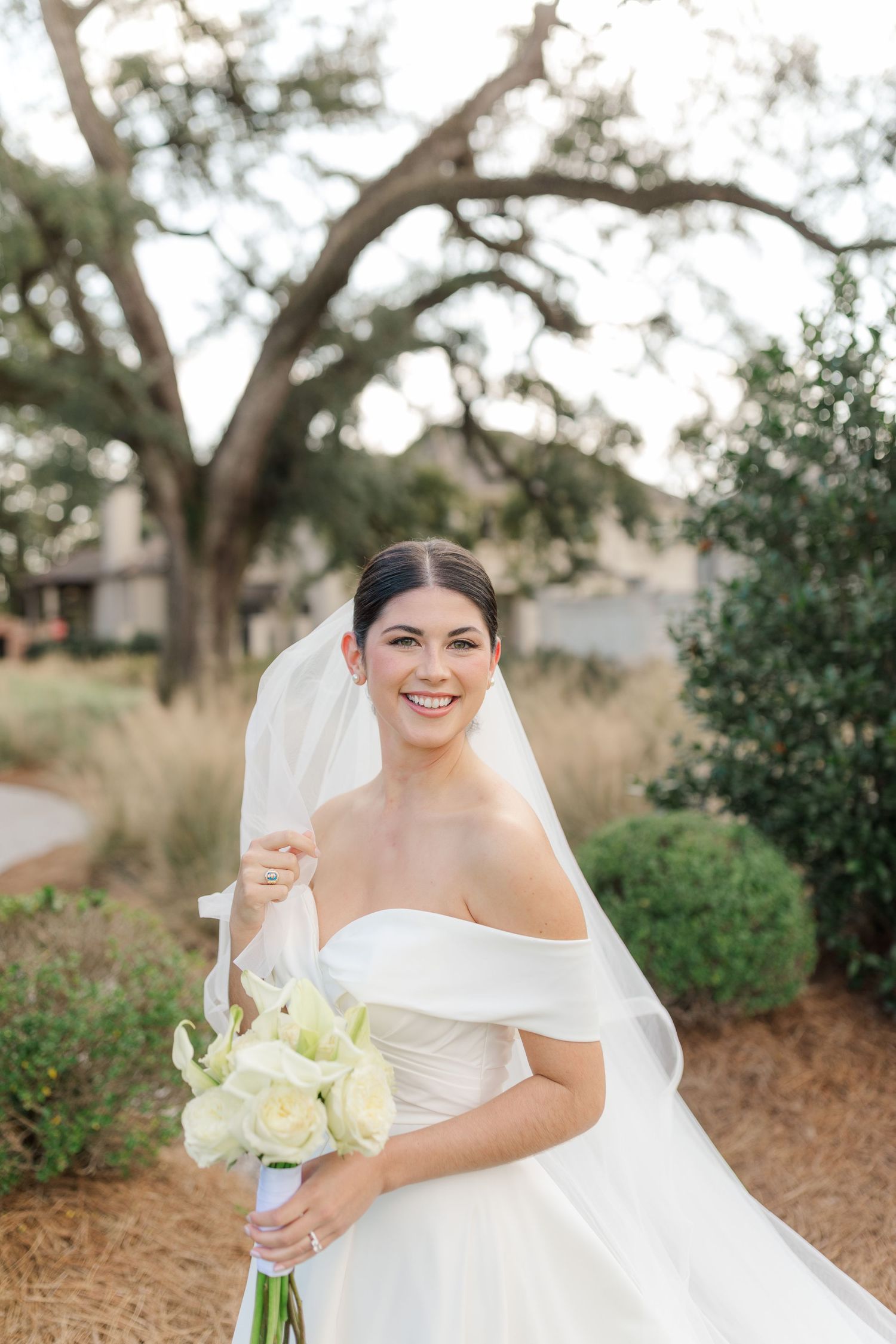 Beautiful bride in off-shoulder white wedding gown holds cream bouquet while posing outdoors under oak trees.