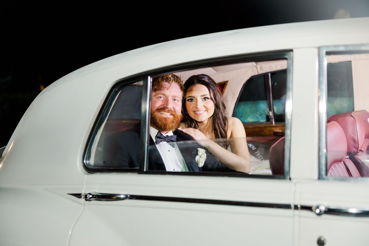Newlyweds smiling through vintage white car windows during their wedding exit.