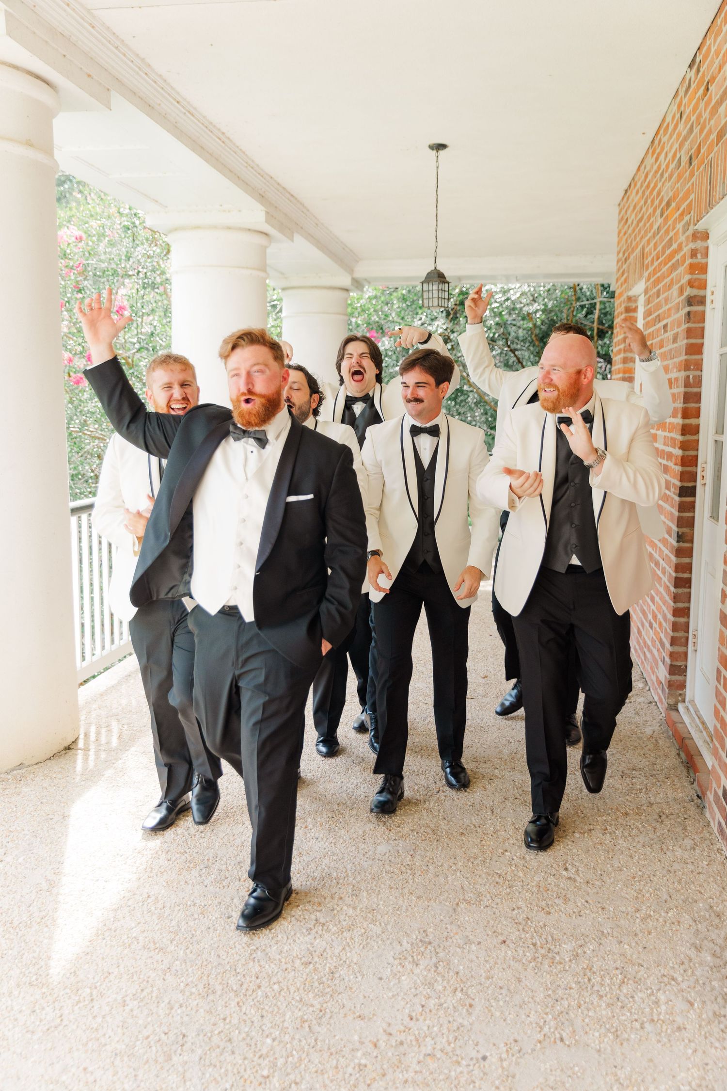 Groomsmen in tuxedos jumping and celebrating in a columned hallway.