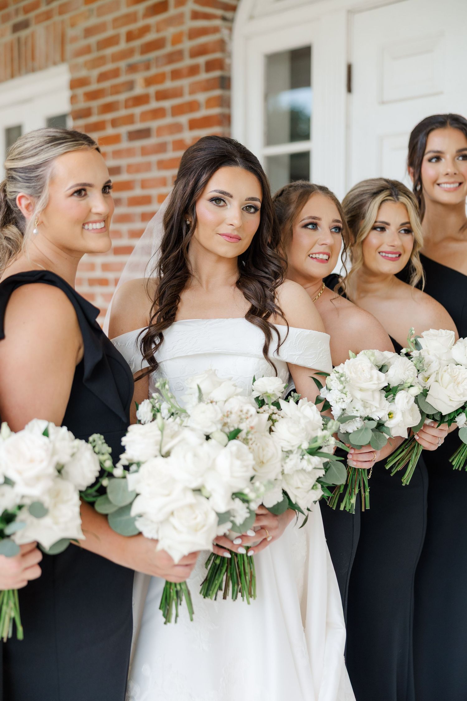 Wedding party poses with white floral bouquets against a brick building backdrop.