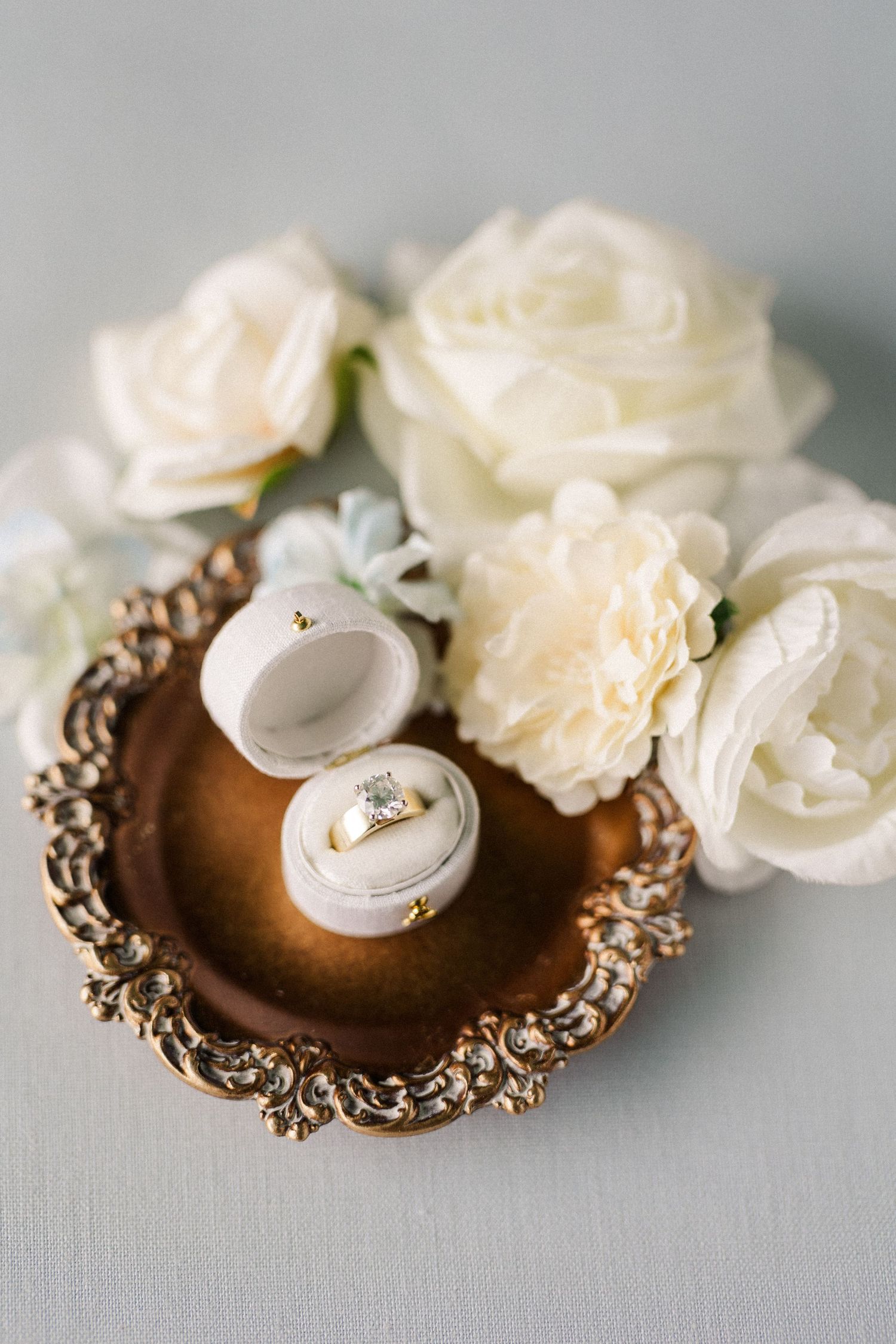 Wedding rings displayed on ornate vintage bronze holders with delicate white roses.