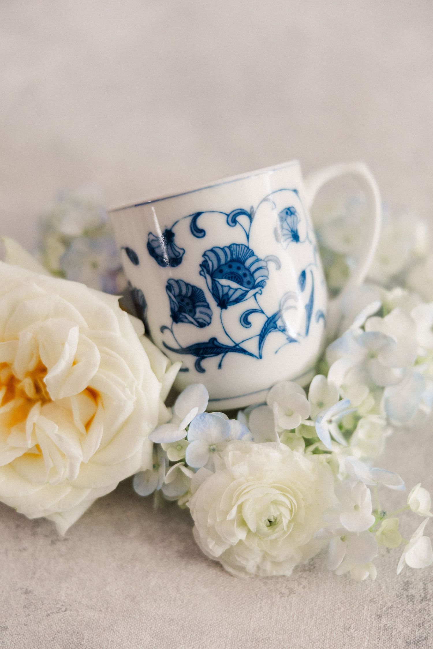 Delicate porcelain teacup with blue floral pattern surrounded by white roses and hydrangeas on light background.