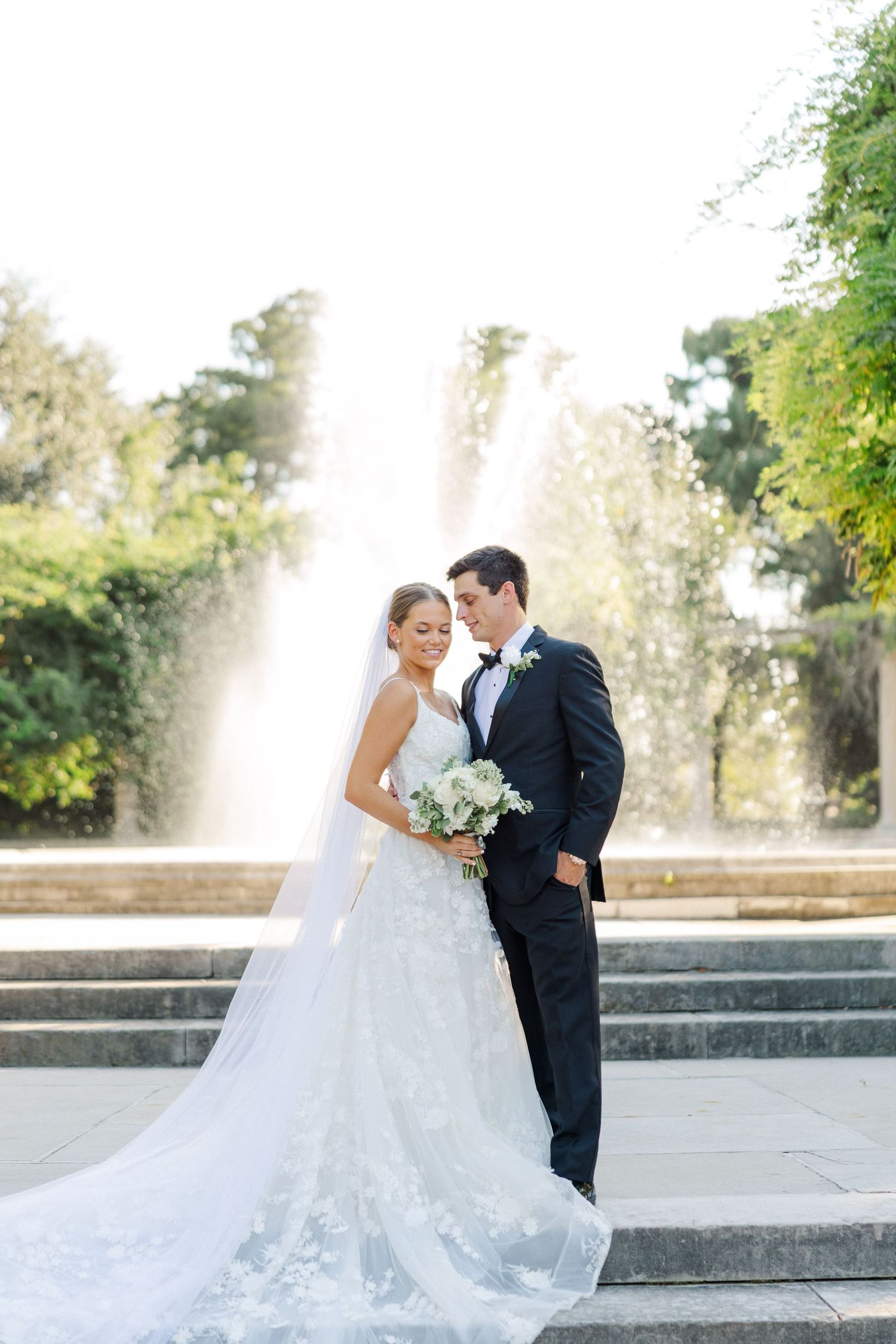 A wedding couple poses on grand stone steps with a white fountain in the background.
