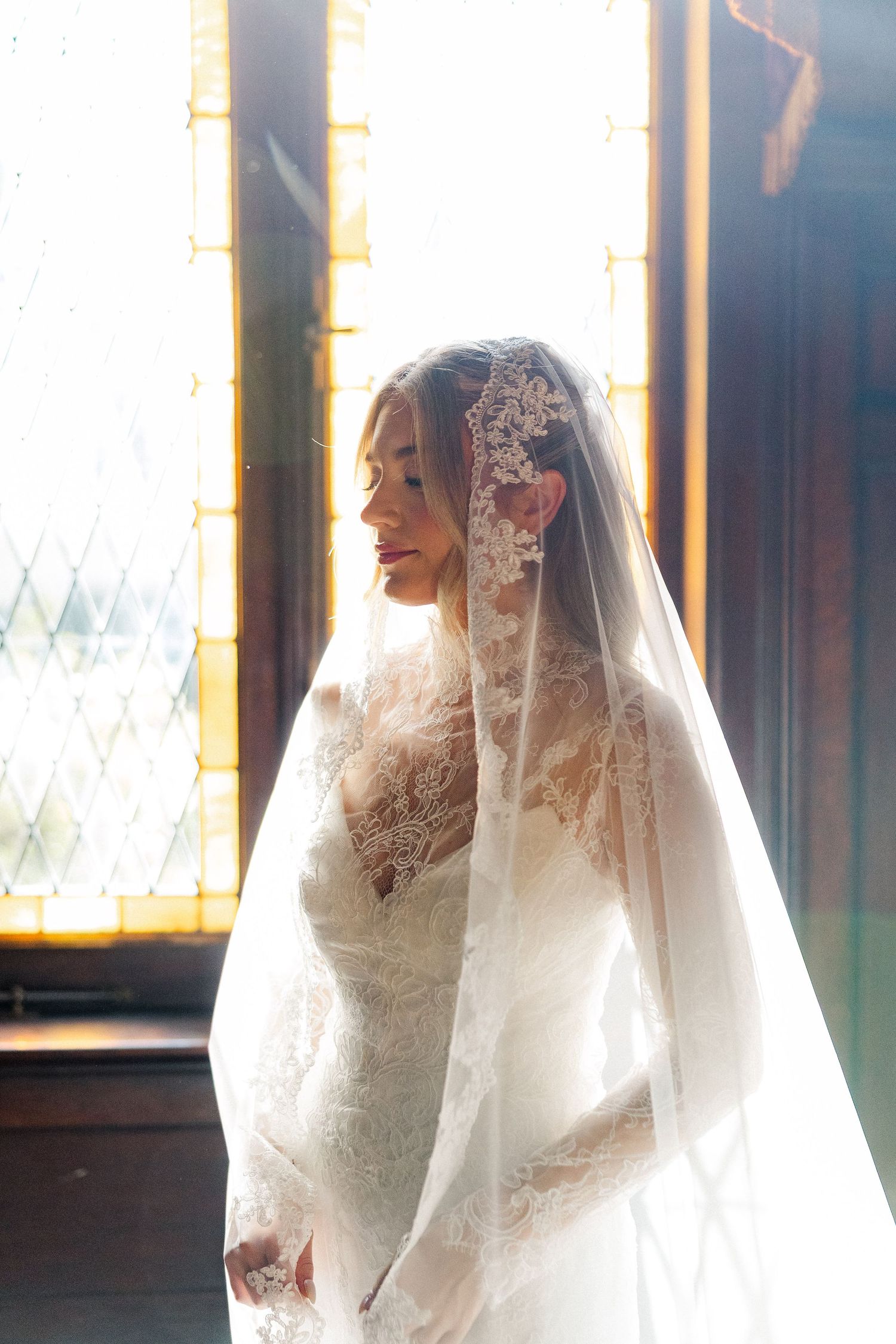 Bride in lace gown and veil illuminated by golden window light in romantic bridal portrait.