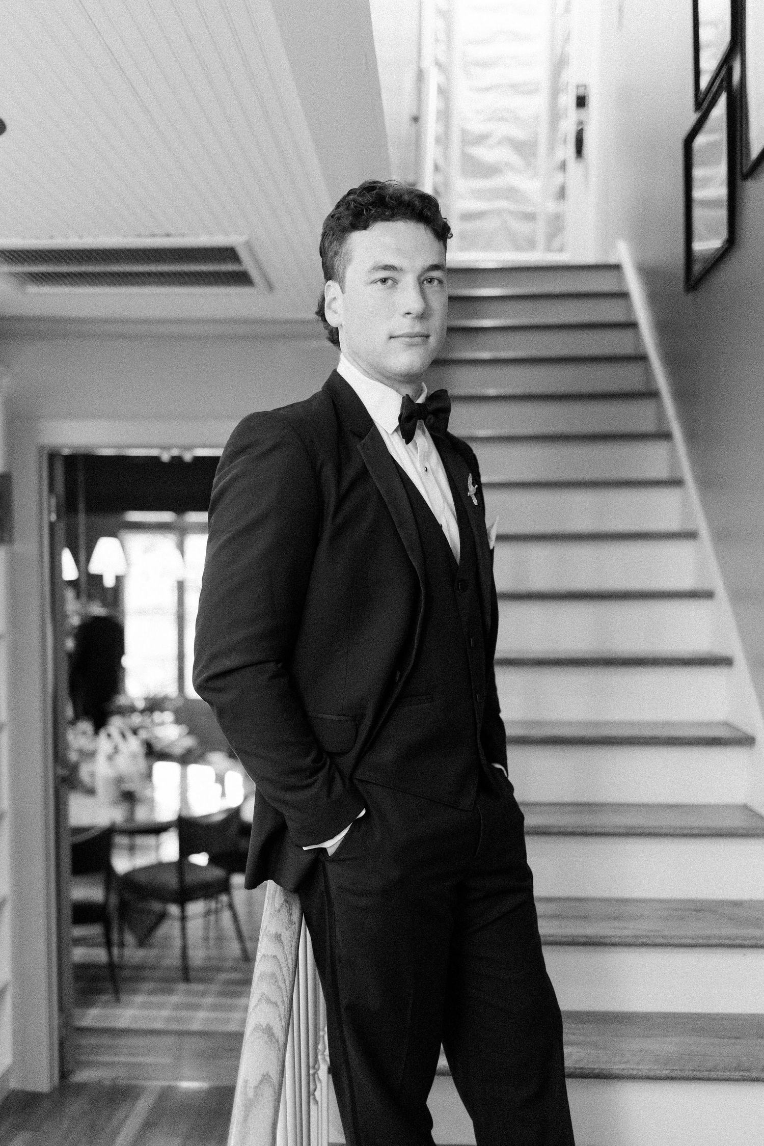 Black and white photos of a groom posing on elegant stairs in a classic tuxedo.