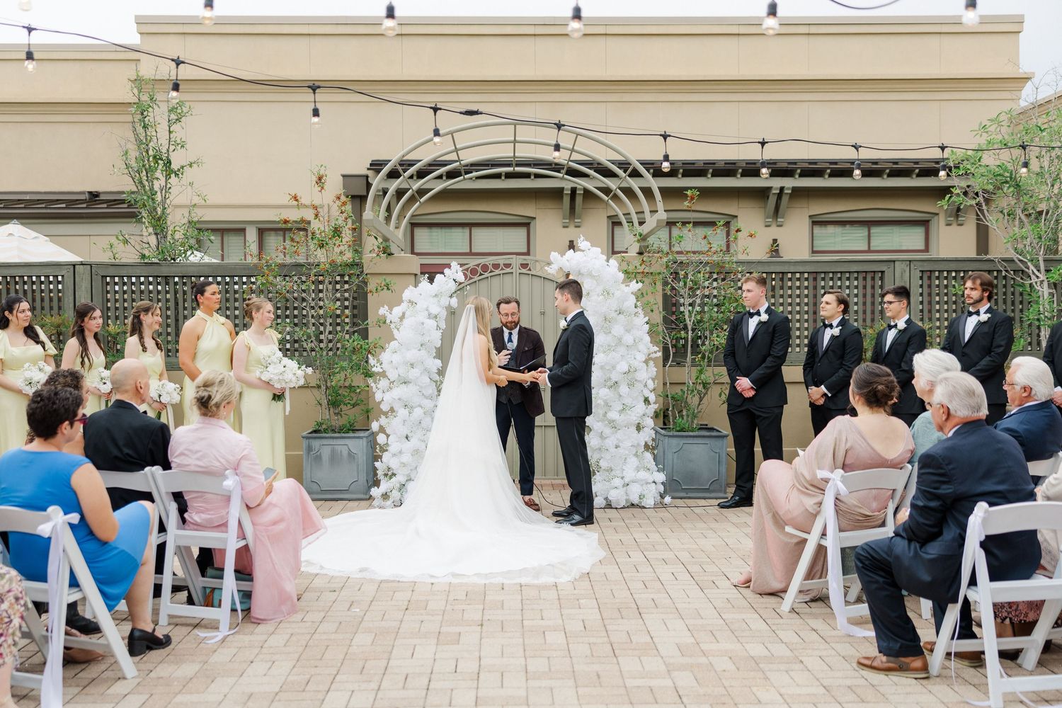 Outdoor wedding ceremony under an arch with white flowers and folding chairs on a patio.