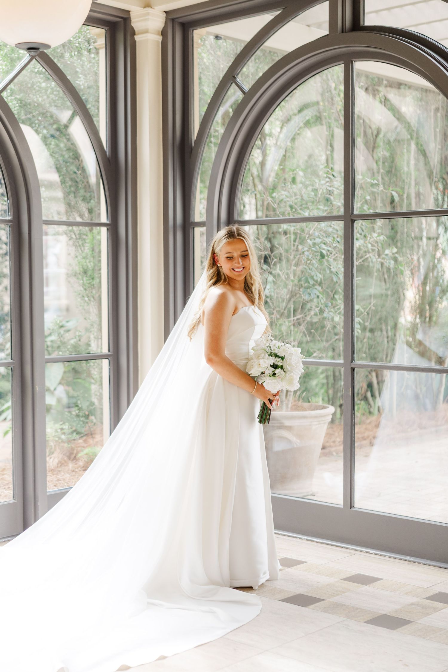 A bride in a flowing white gown and veil poses elegantly in front of large arched windows.