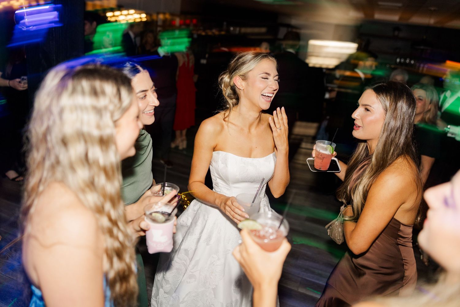 Wedding reception party guests dancing under colorful lights in a dimly lit venue.
