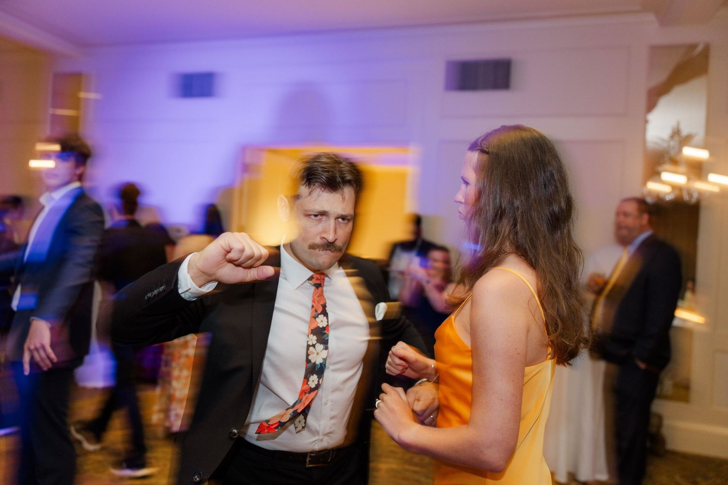 Wedding guests dance and celebrate on a reception dance floor with purple lighting.