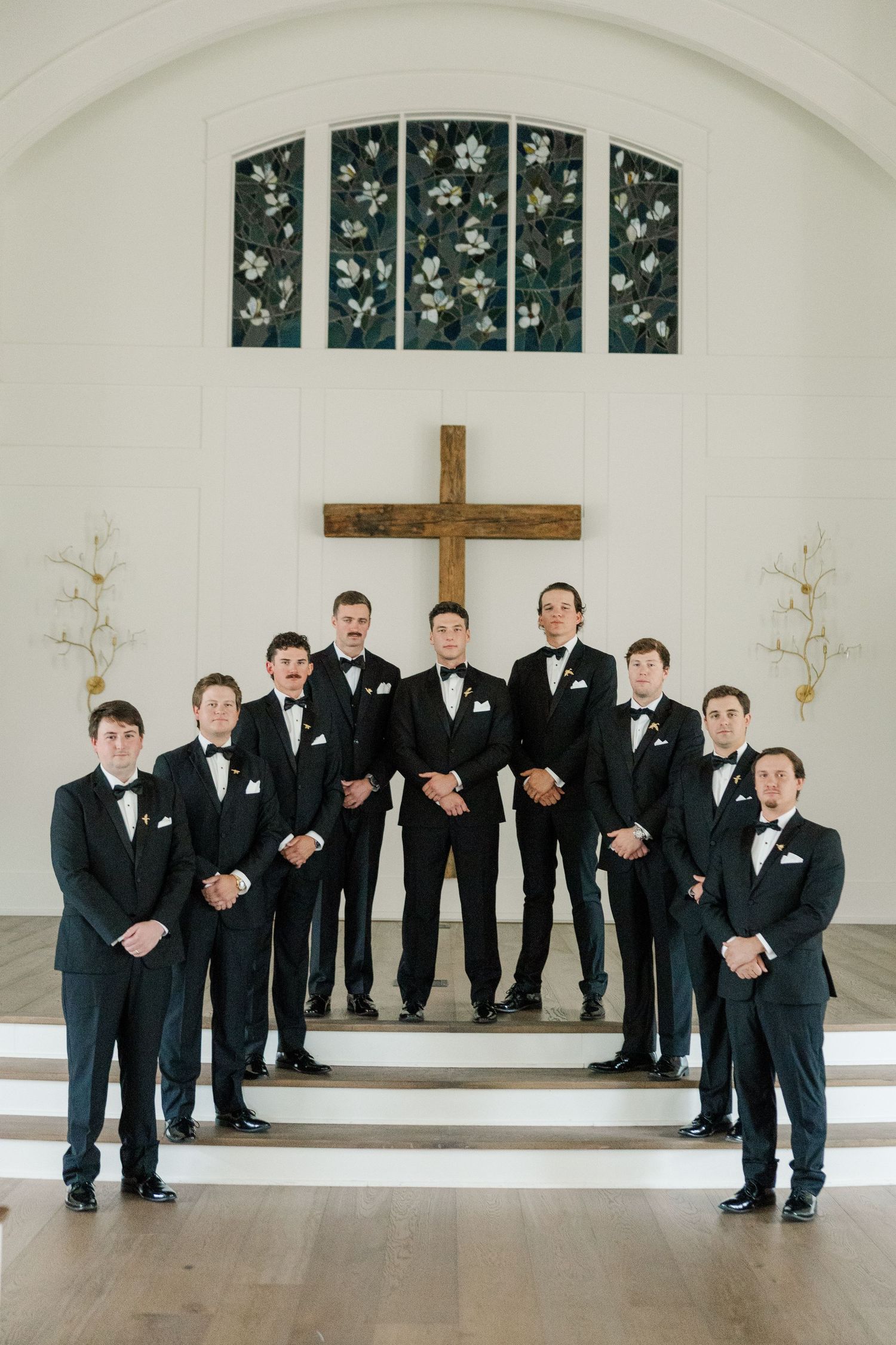 Groomsmen in black tuxedos stand together in a bright white church interior with a wooden cross on the wall.