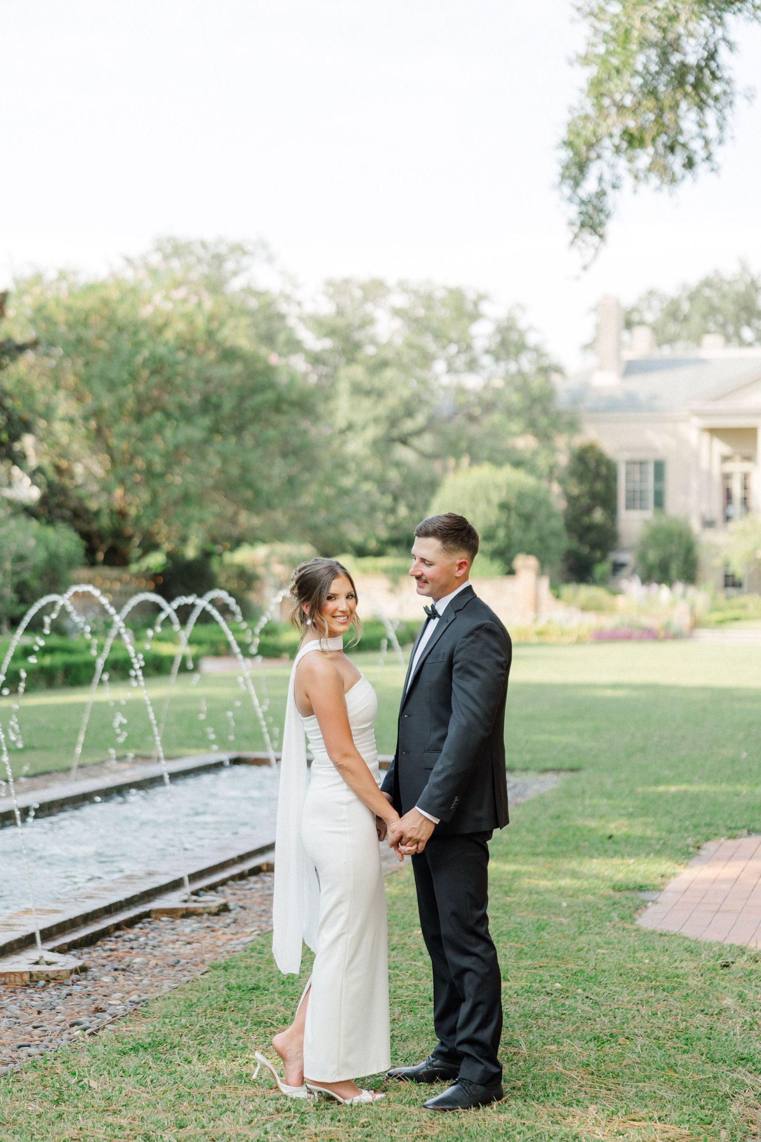 A couple poses by a water fountain feature in a garden setting wearing elegant wedding attire.