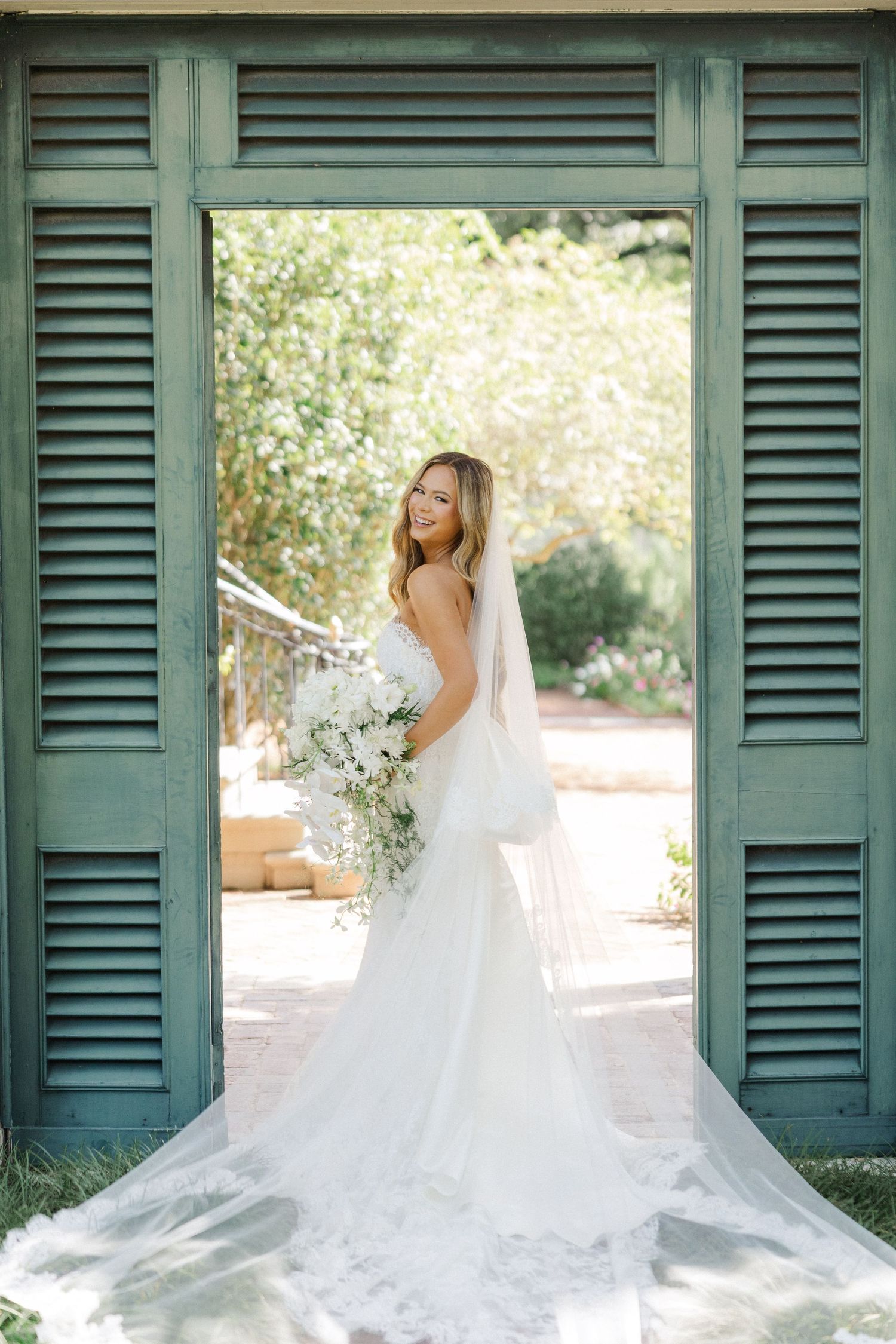 Classic bridal portrait framed by teal shutters showcasing a flowing white wedding gown with long train.