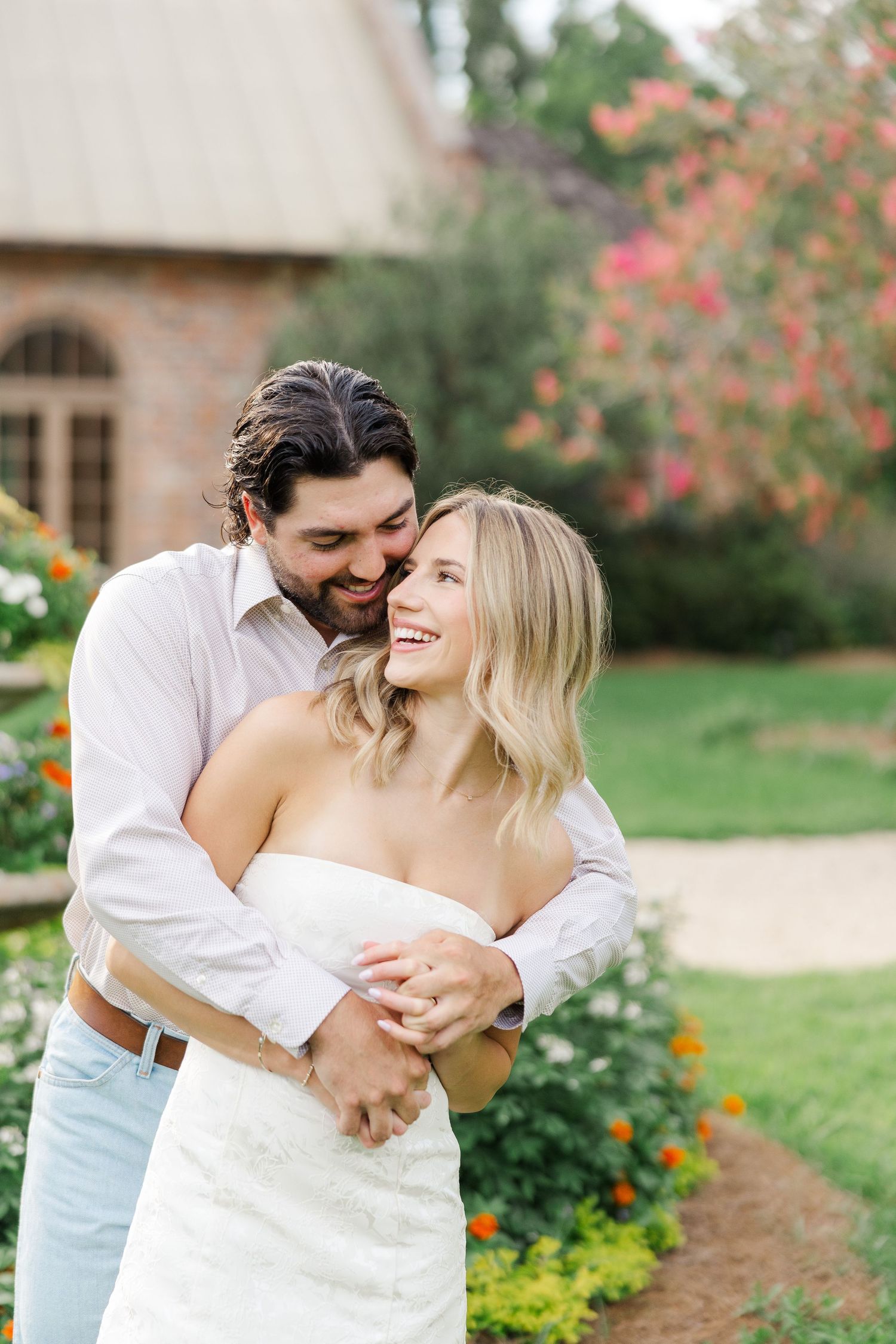 A romantic couple embraces in a garden setting with blooming flowers and a brick building in the background.