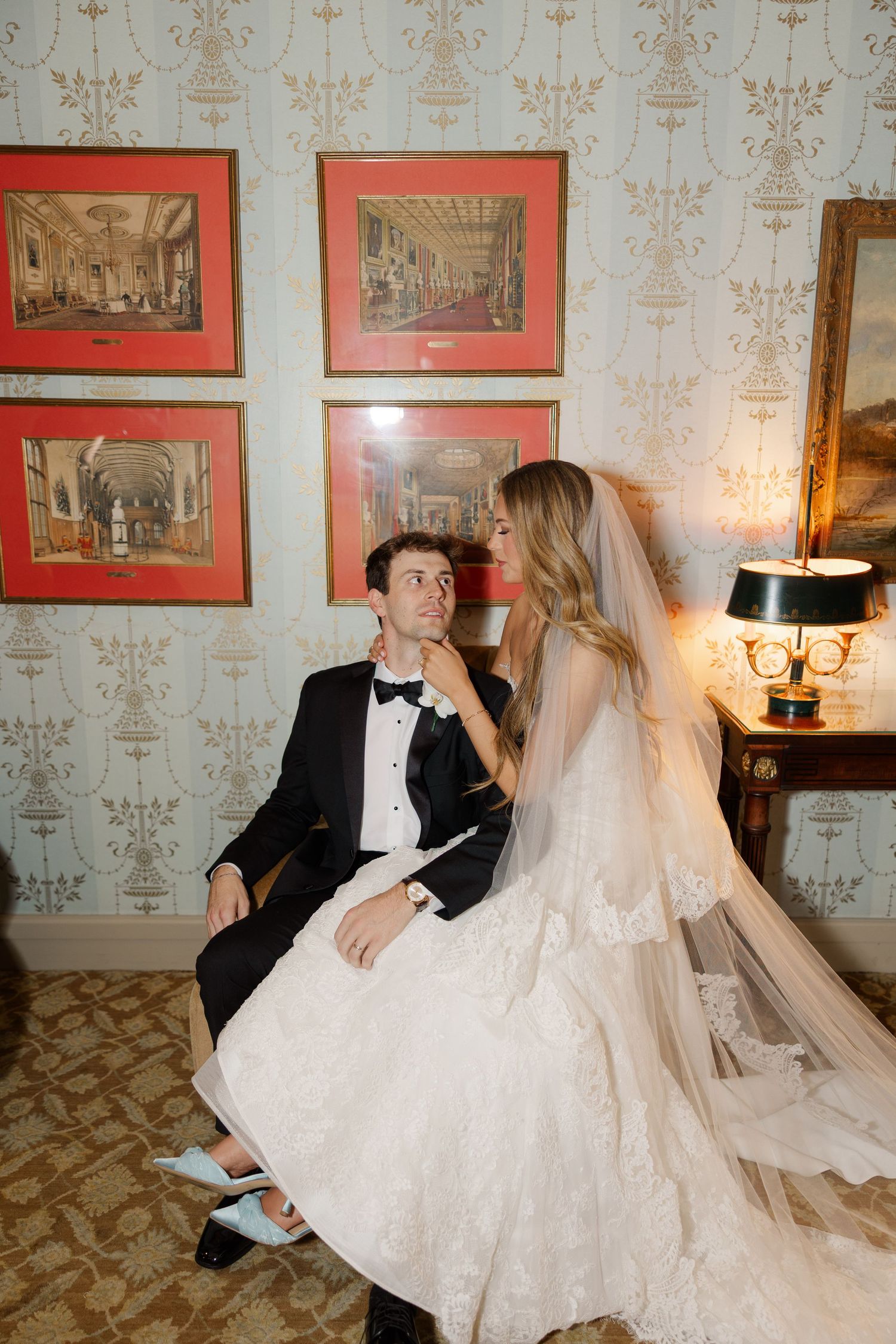 Romantic wedding couple poses together on vintage furniture in an elegant room with patterned wallpaper.