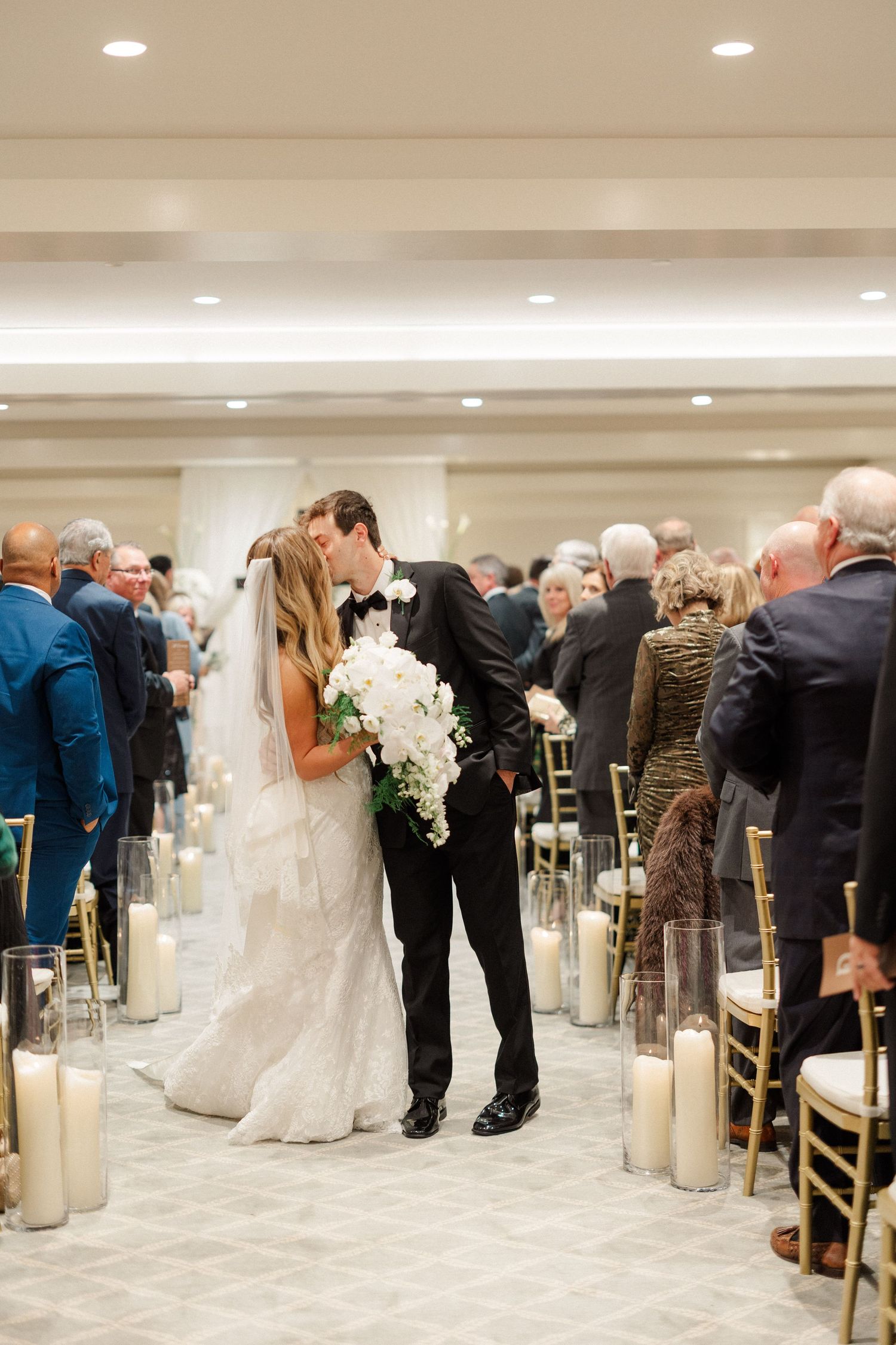 Wedding reception in an elegant white room with candles lining the aisle and gold chairs.