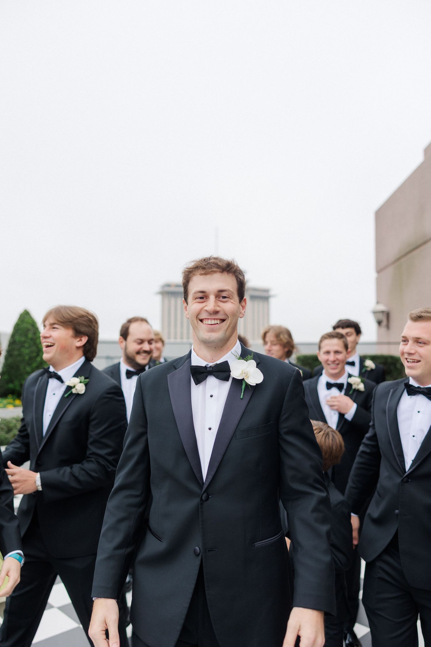 A group of groomsmen in black tuxedos and bow ties share a laugh together at a wedding celebration.