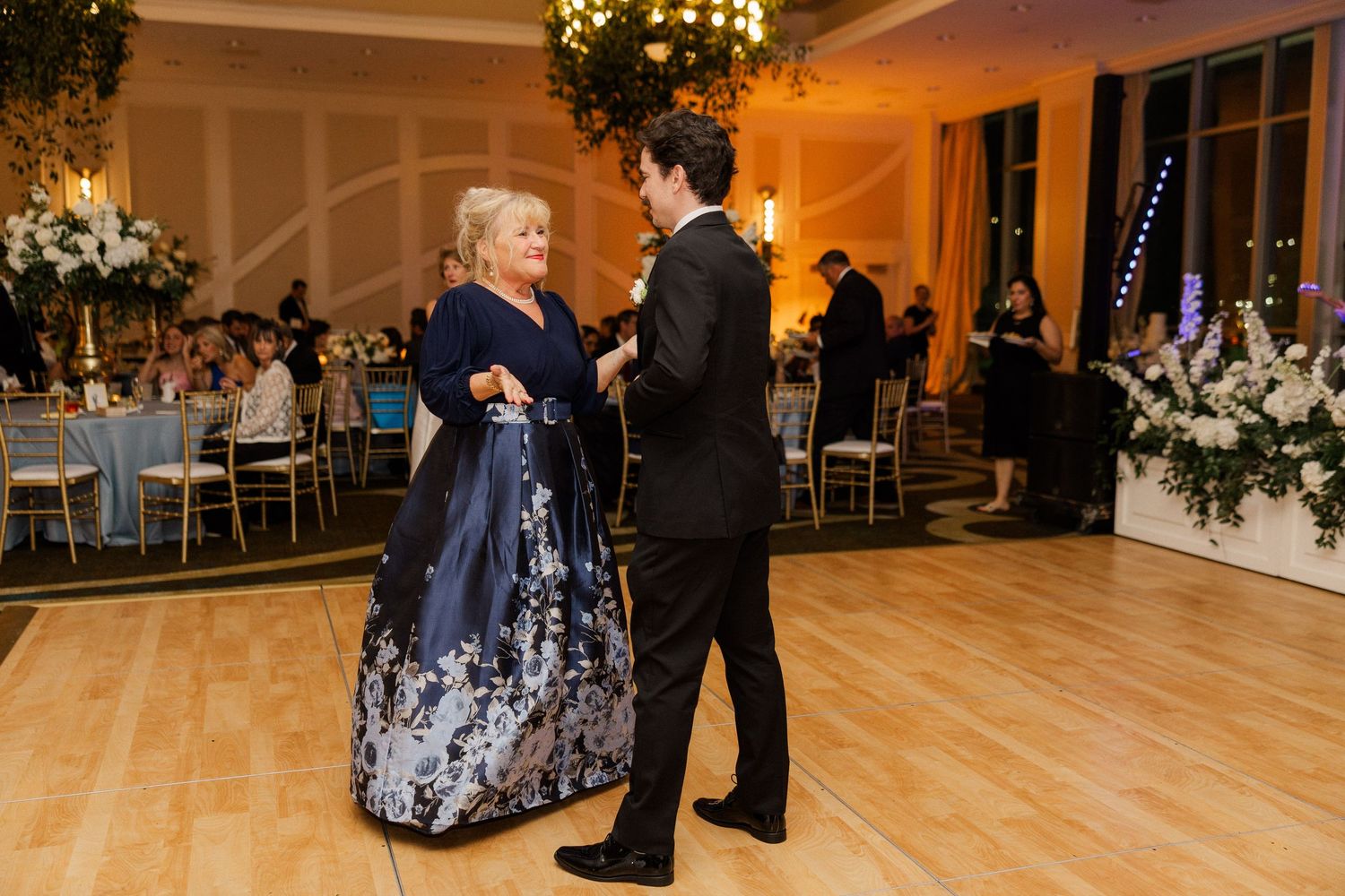 Groom in black tuxedo shares mother-son dance in elegant ballroom reception with floral centerpieces.