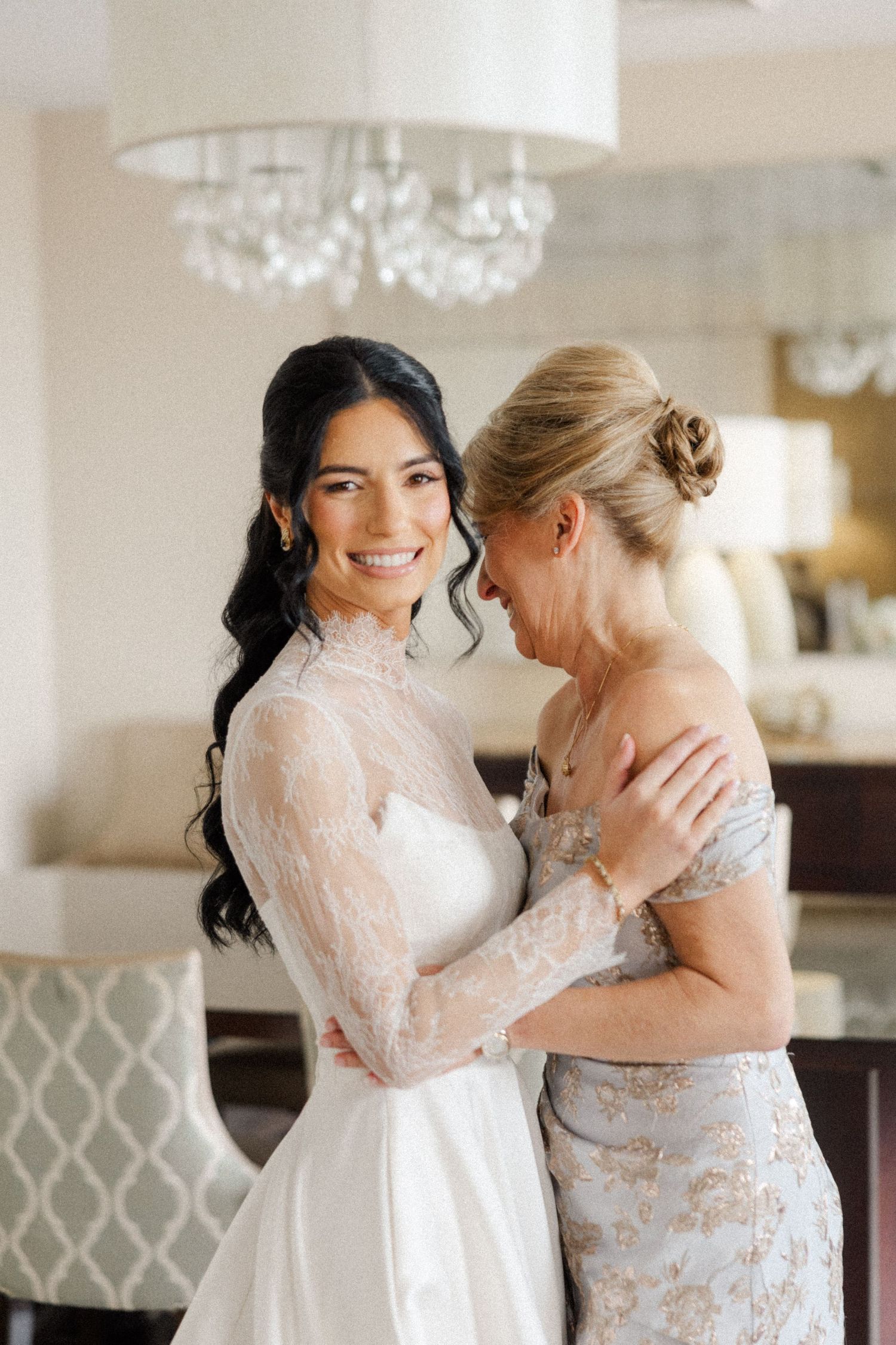 Bride in lace long-sleeve wedding gown smiles with older woman in silver dress during bridal preparations.