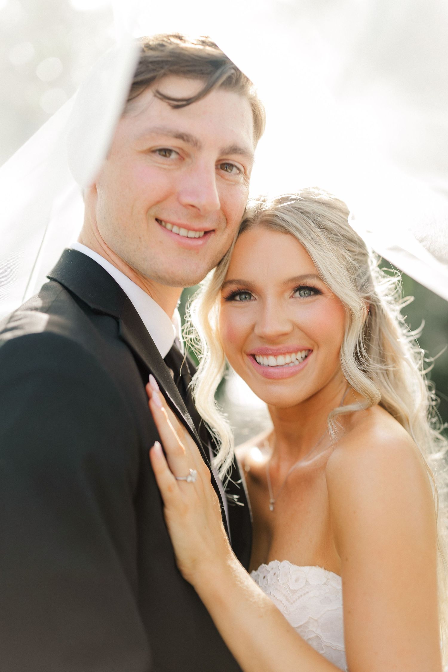 Smiling bride and groom pose closely together under a veil in soft golden sunlight on their wedding day.