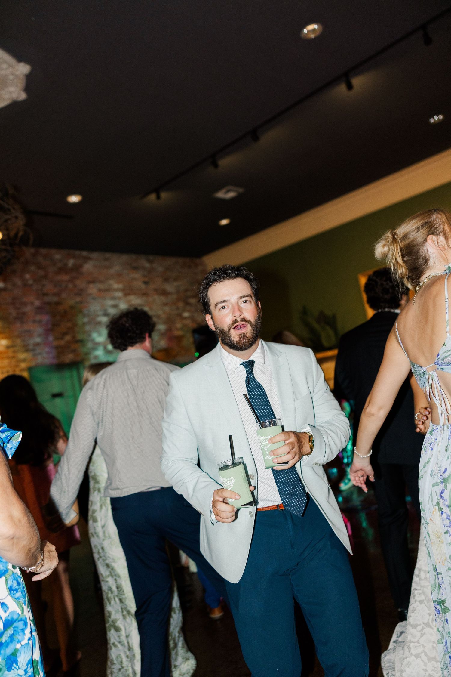 Wedding guest in light suit holding cocktail on crowded dance floor at reception.