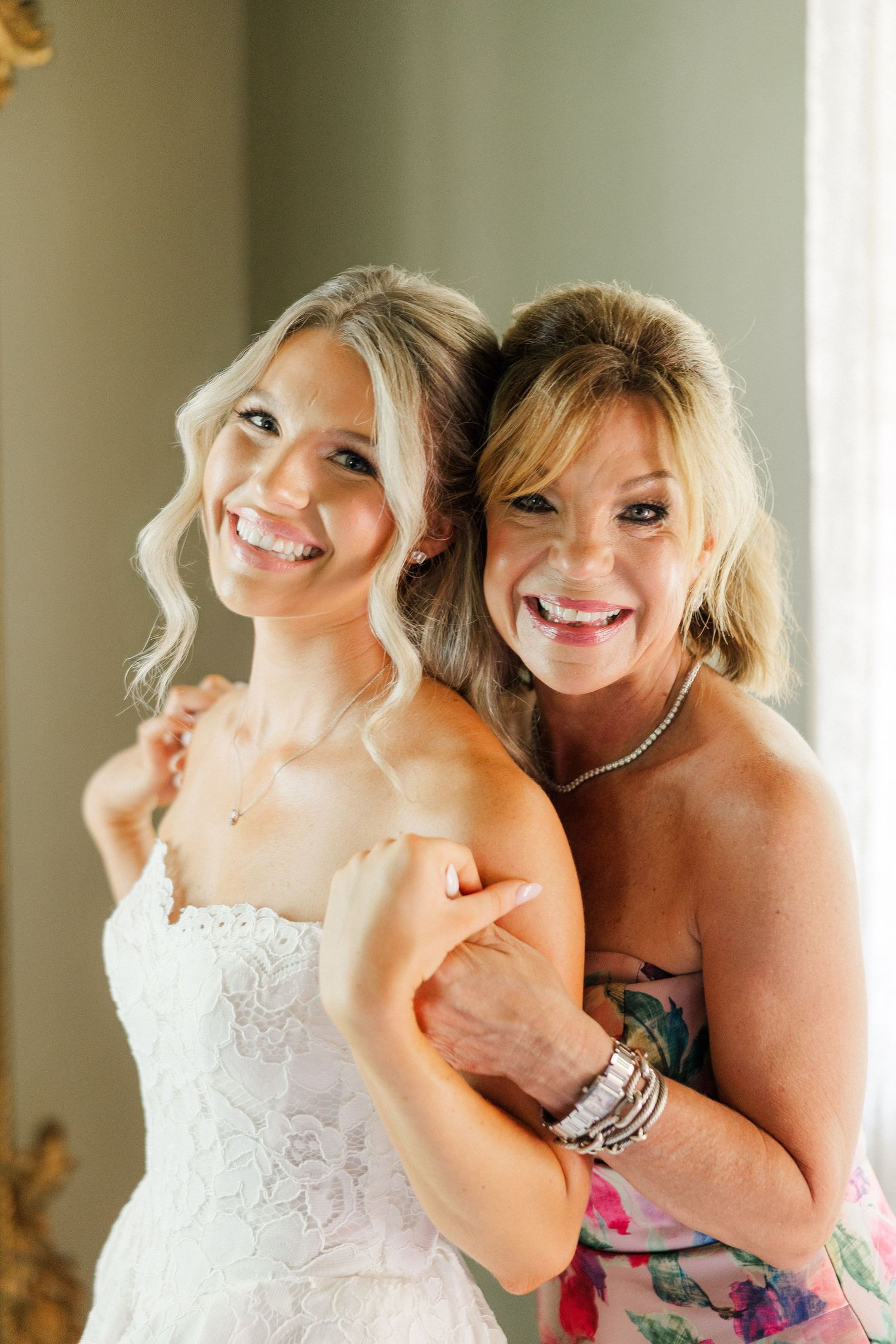 Smiling bride in strapless lace gown embraced by older woman in floral dress during wedding preparations.