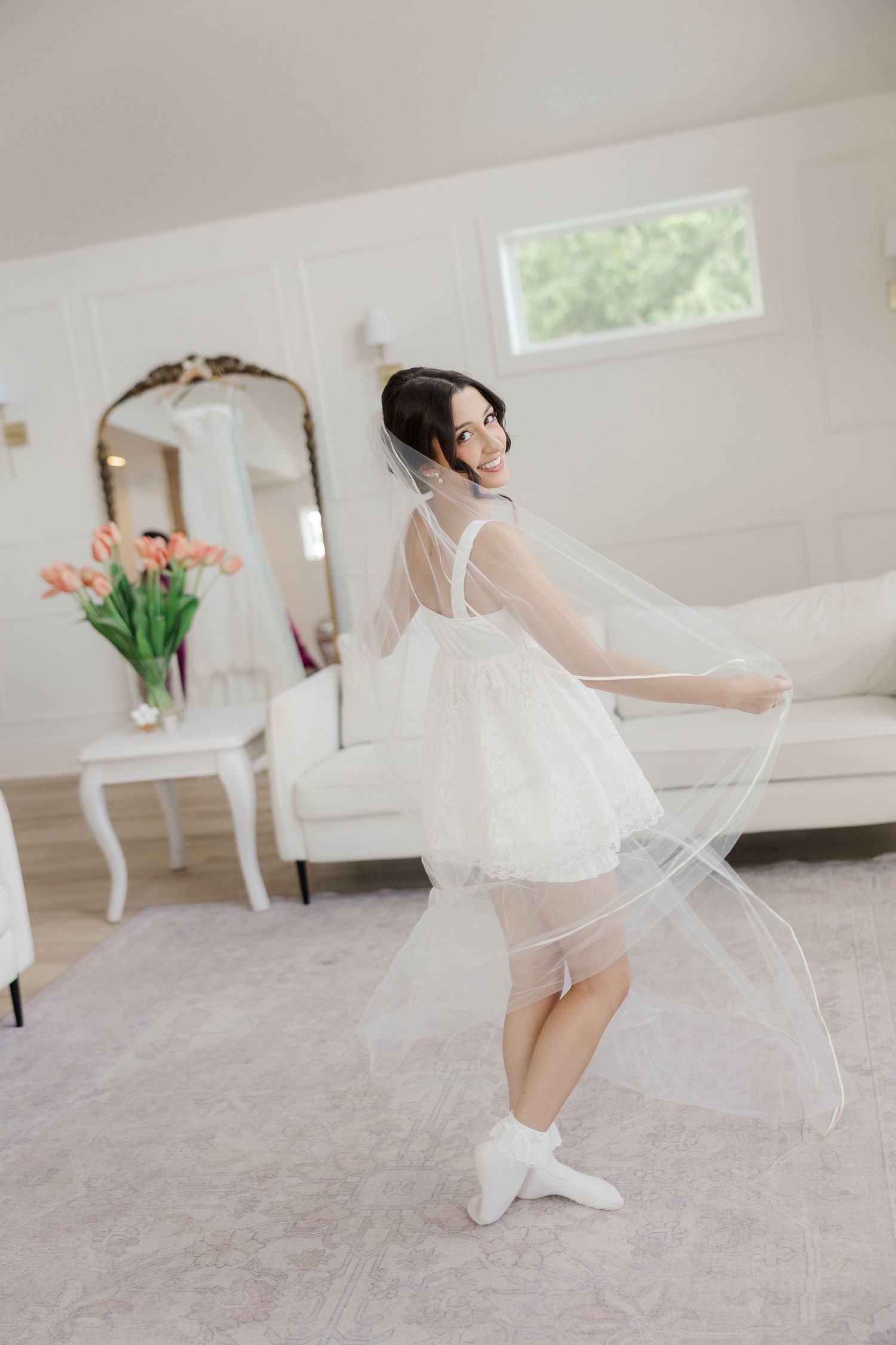 Smiling bride in short white dress and veil twirling in a bright white bridal suite before her wedding.