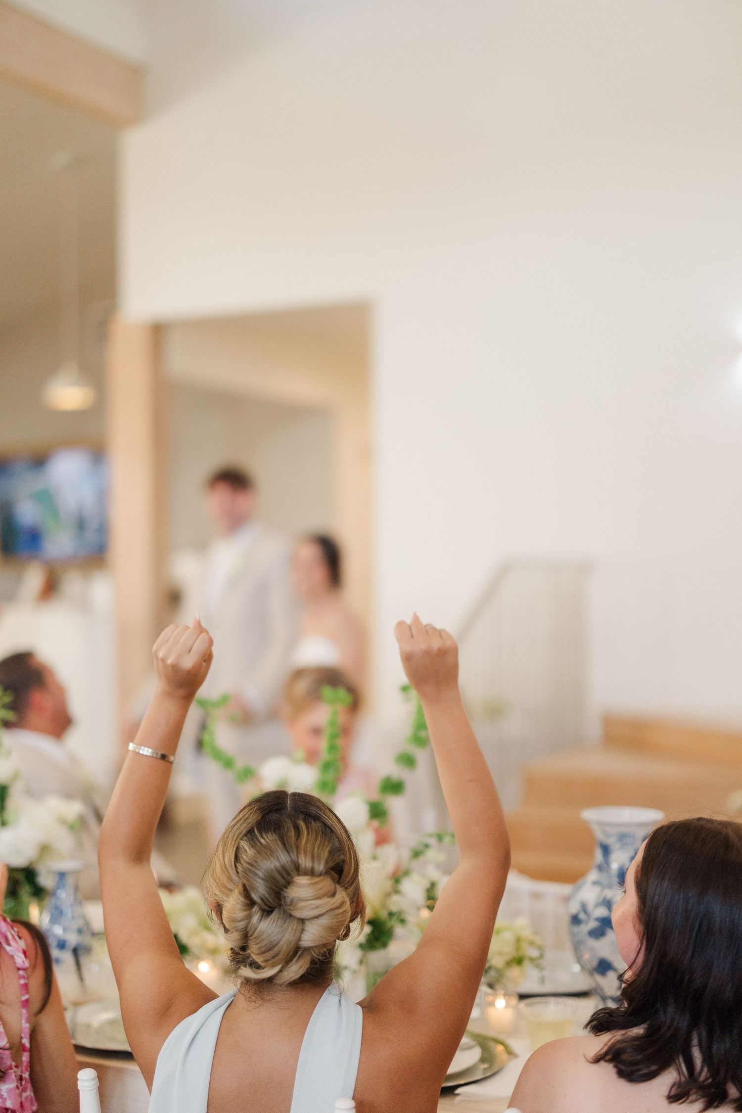 Wedding guest raises fists in celebration as bride and groom enter the reception hall in background.