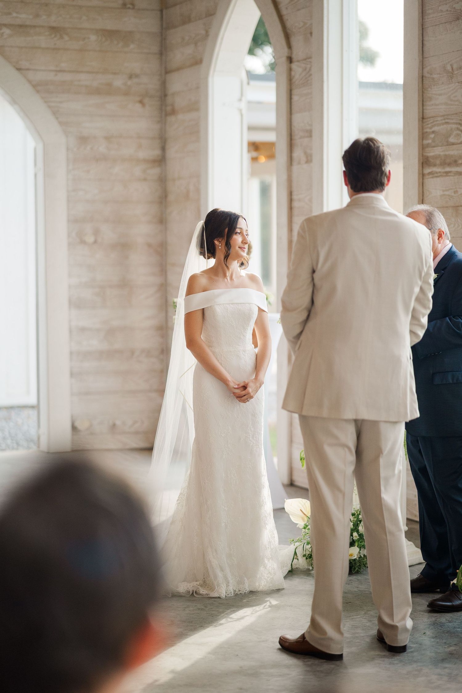Smiling bride in off-shoulder white gown and veil stands at altar facing groom in beige suit during wedding ceremony.