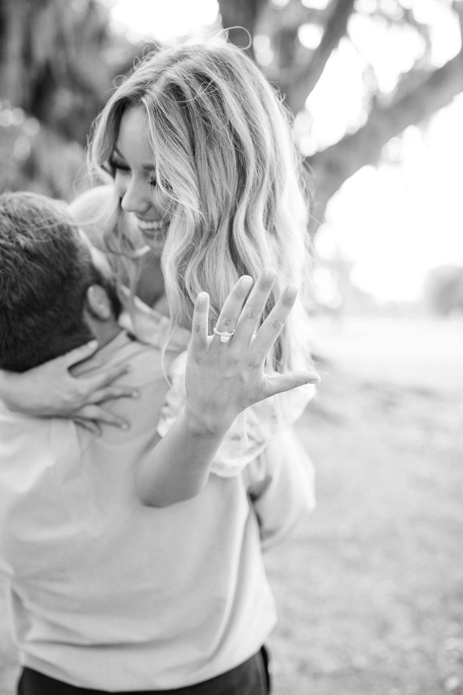 A romantic black and white engagement photo showing a joyful couple embracing outdoors with a sparkling ring display.