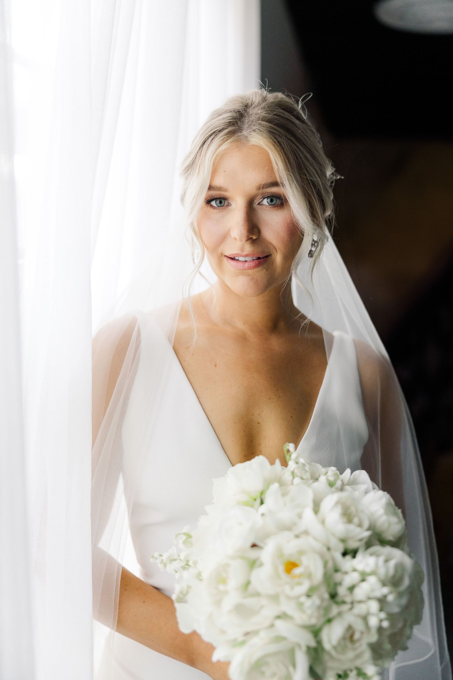 Elegant bride in white V-neck dress holds a lush bouquet of white peonies against sunlit window.