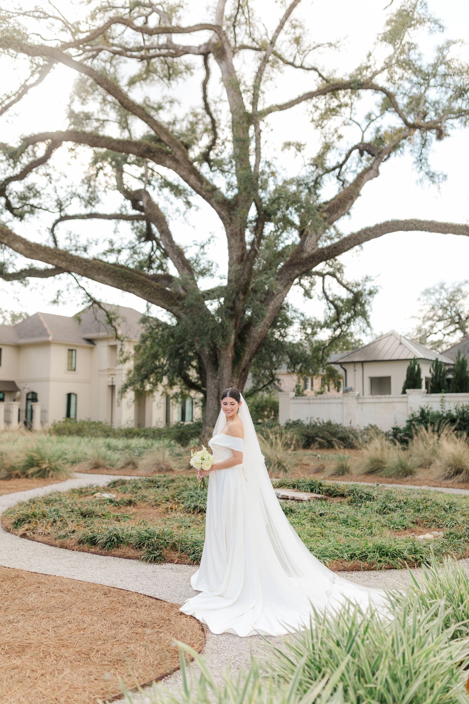 A bride in a flowing white gown poses on a winding garden path beneath a majestic oak tree.