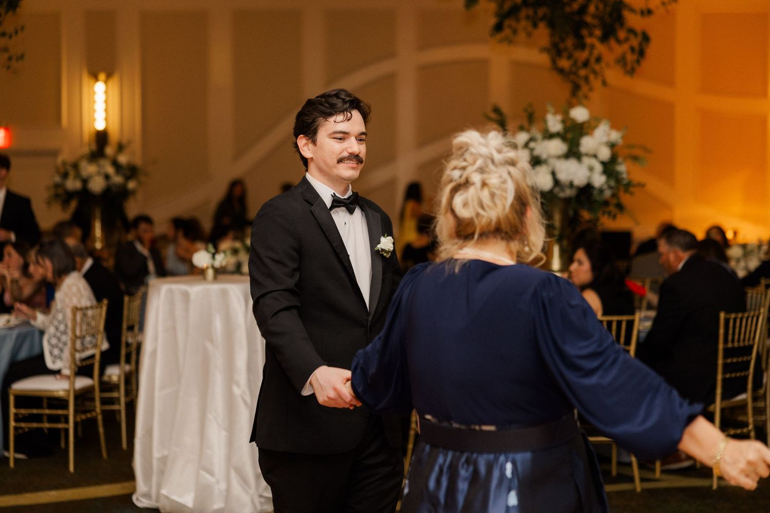 Groom in black tuxedo shares dance with woman in navy dress at elegant wedding reception.