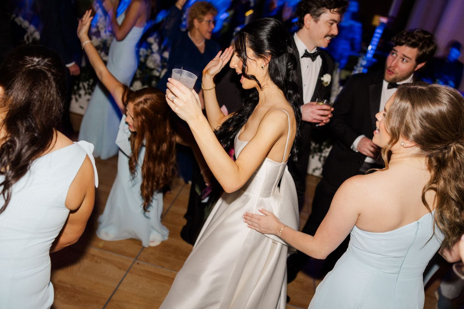 Bride in white satin gown dances and laughs with guests at wedding reception.