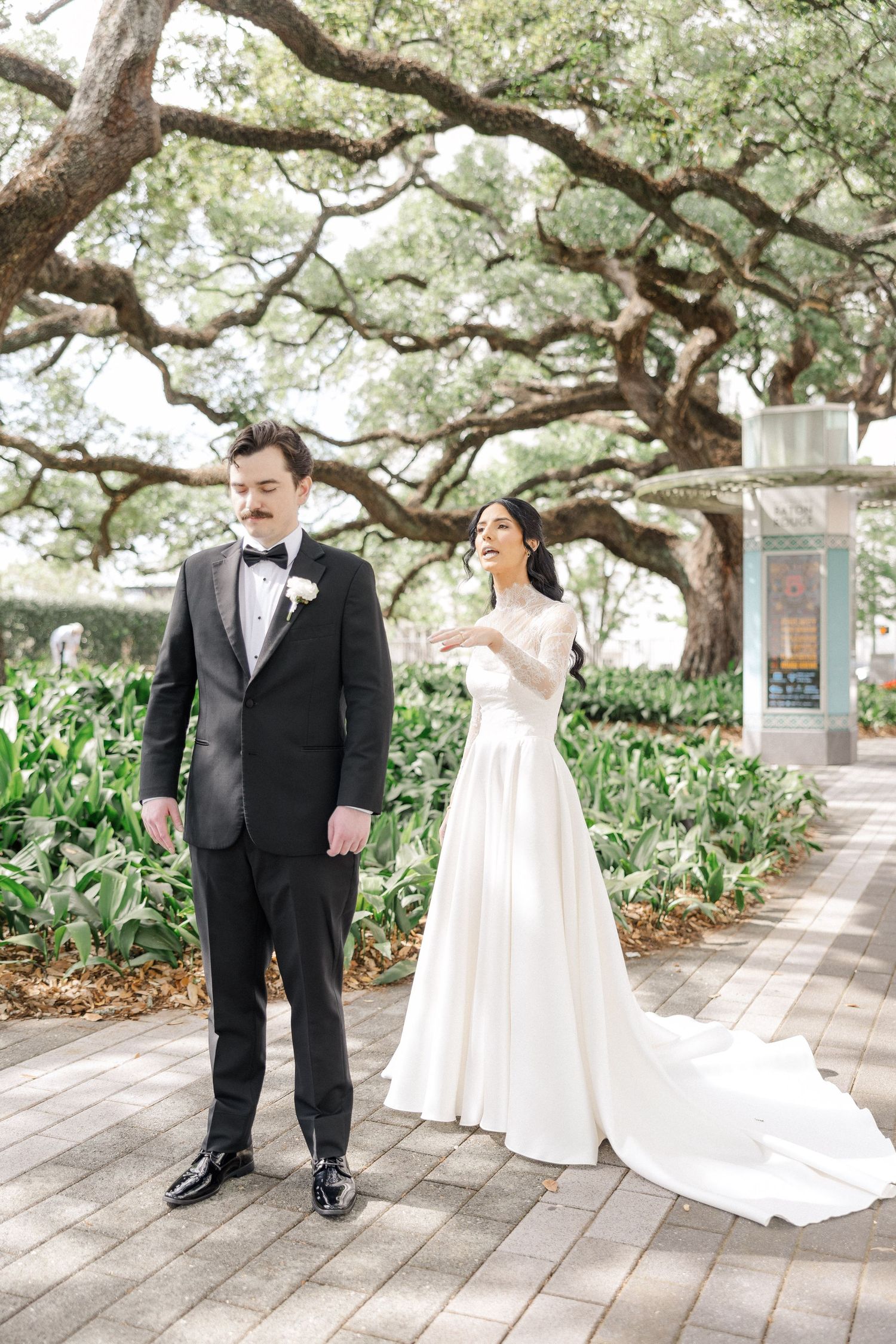 Bride in white gown laughs behind groom in black tuxedo during first look under oak tree.