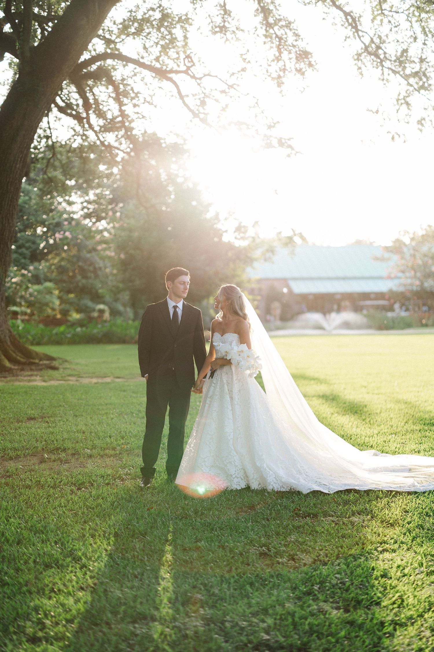 Bride and groom walk hand in hand on lush green lawn bathed in golden sunset light.