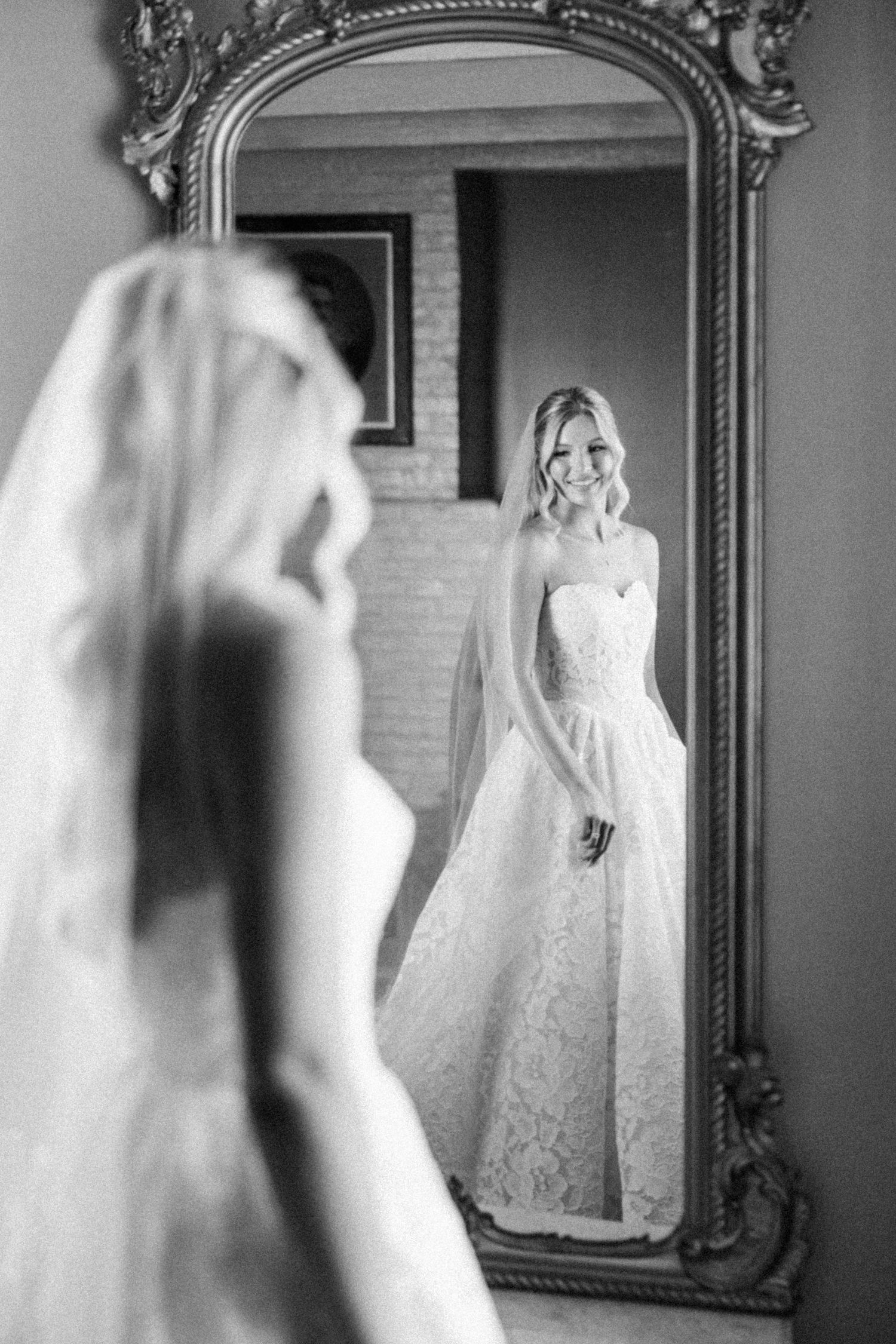 Black and white photo of a smiling bride in lace gown and veil admiring her reflection in an ornate gold mirror.