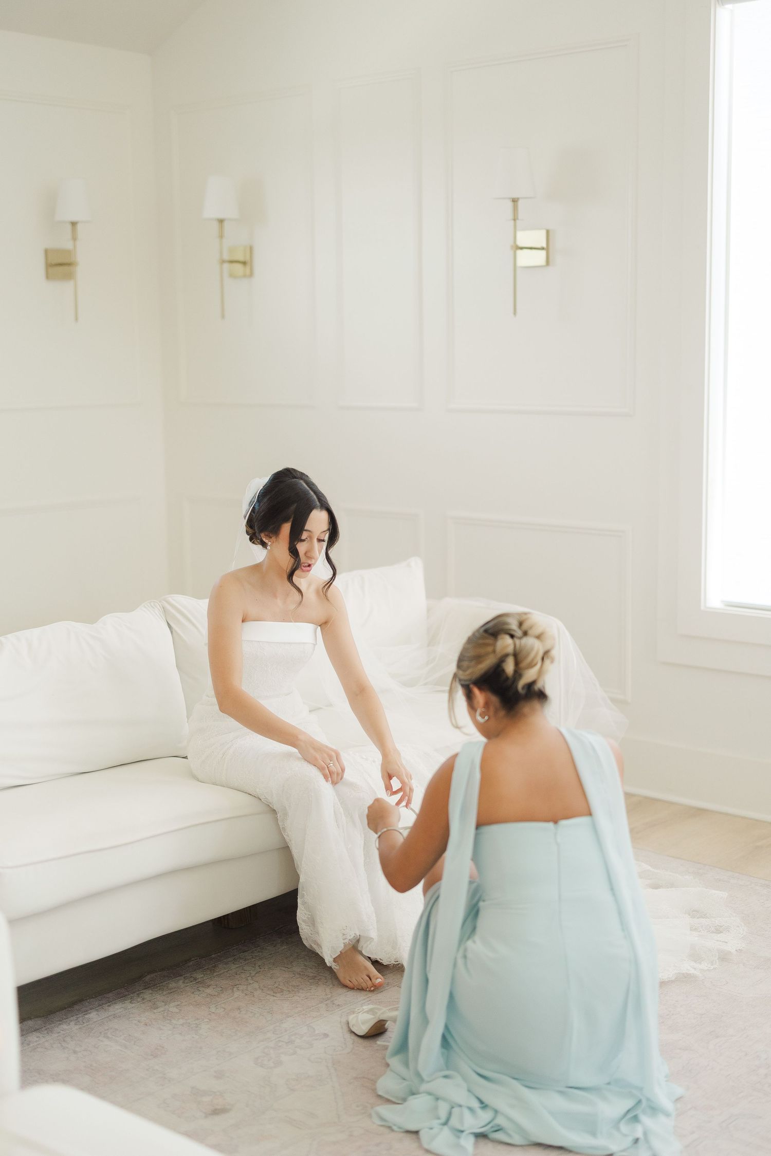 Bridesmaid in light blue dress helping bride with shoes while seated on white sofa in elegant bridal suite.