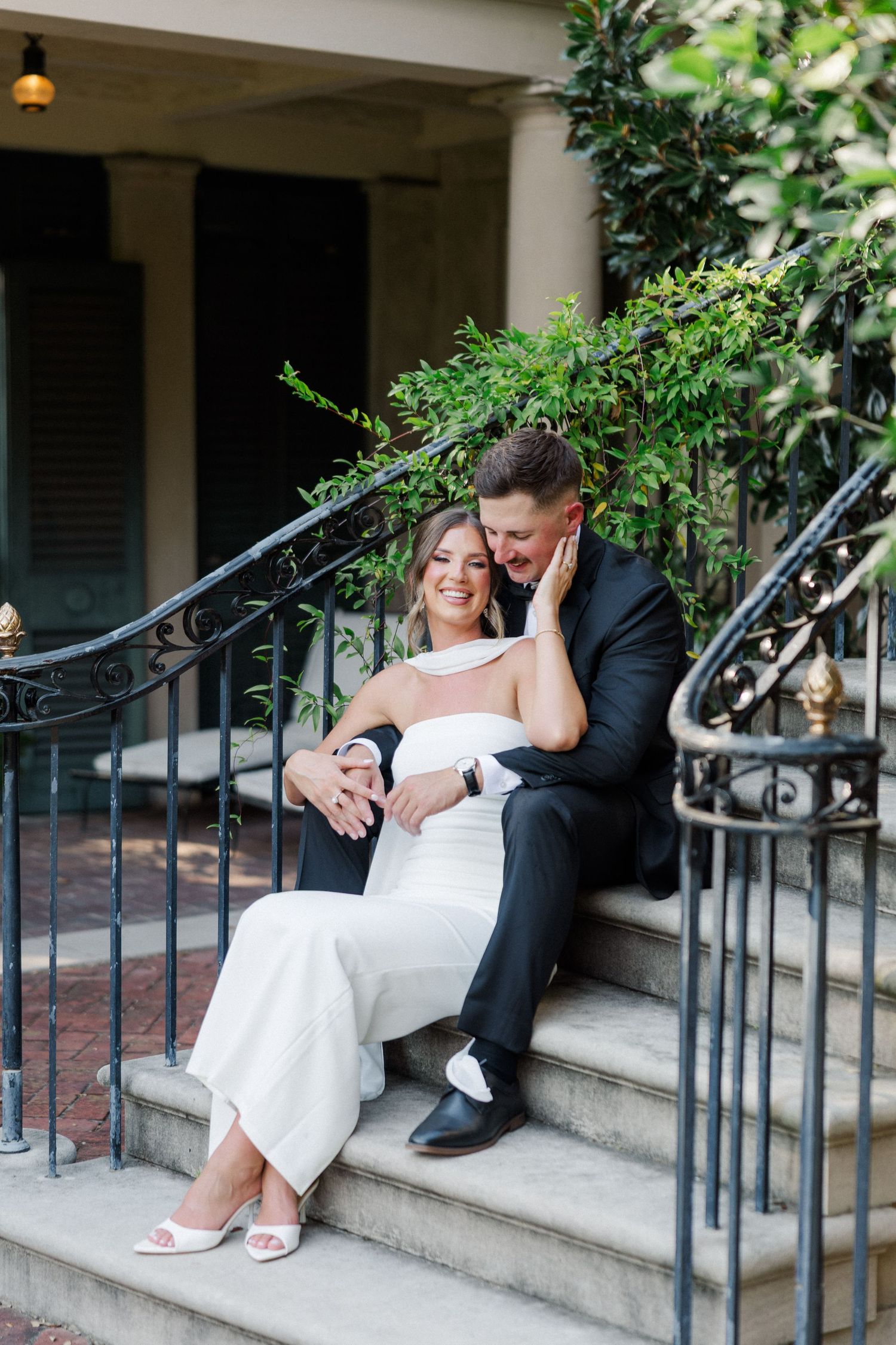 Wedding couple sits romantically on stone steps of a columned building with wrought iron railings.