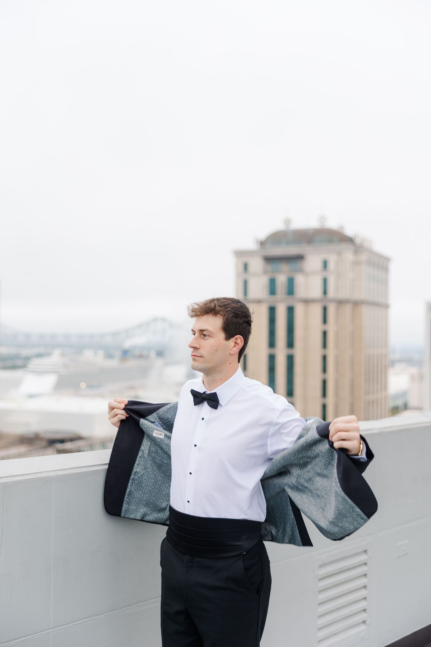 A groom in a tuxedo adjusts his jacket on a rooftop with city views.