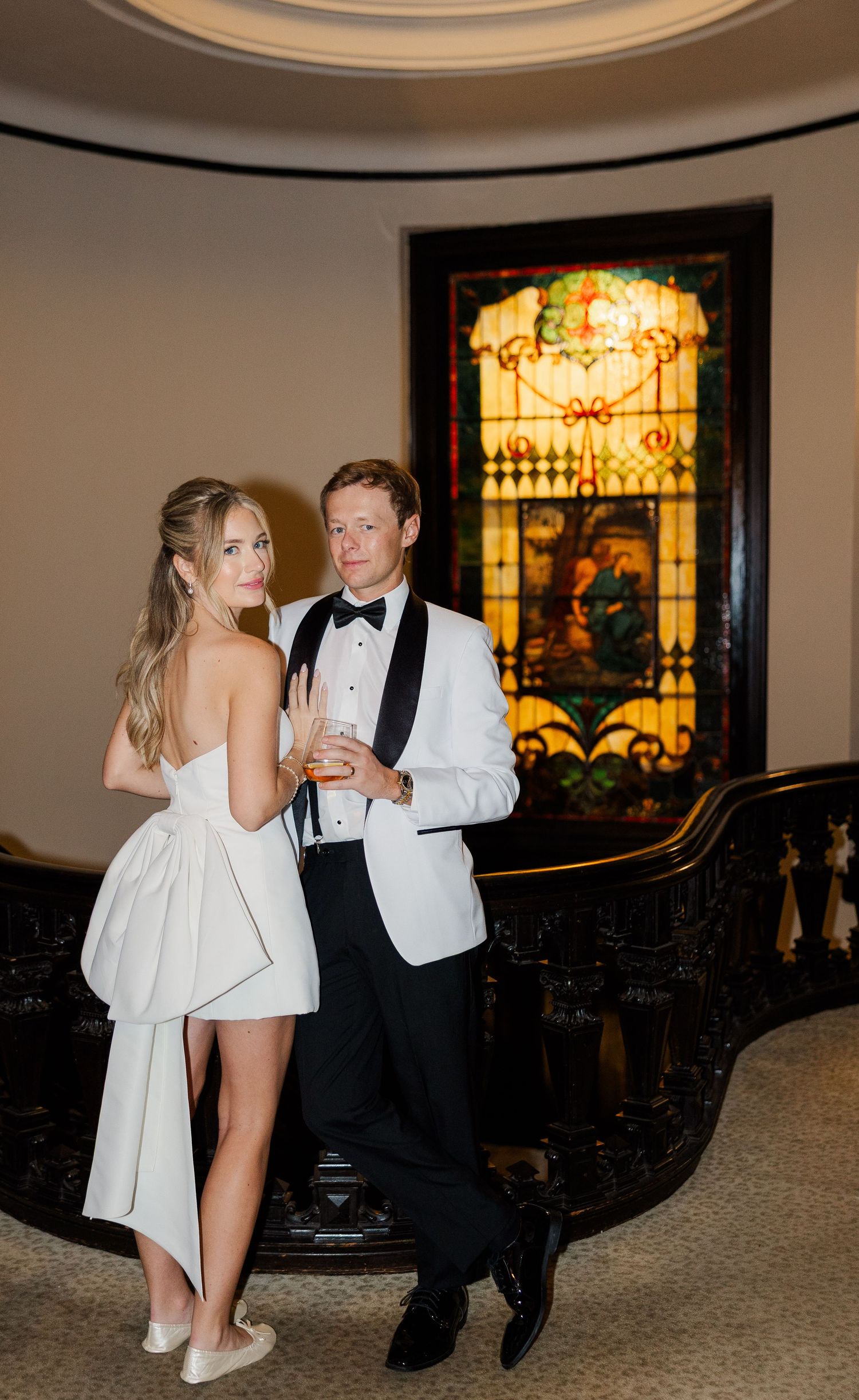 A couple in formal attire poses in front of an elegant stained glass window at an upscale venue.