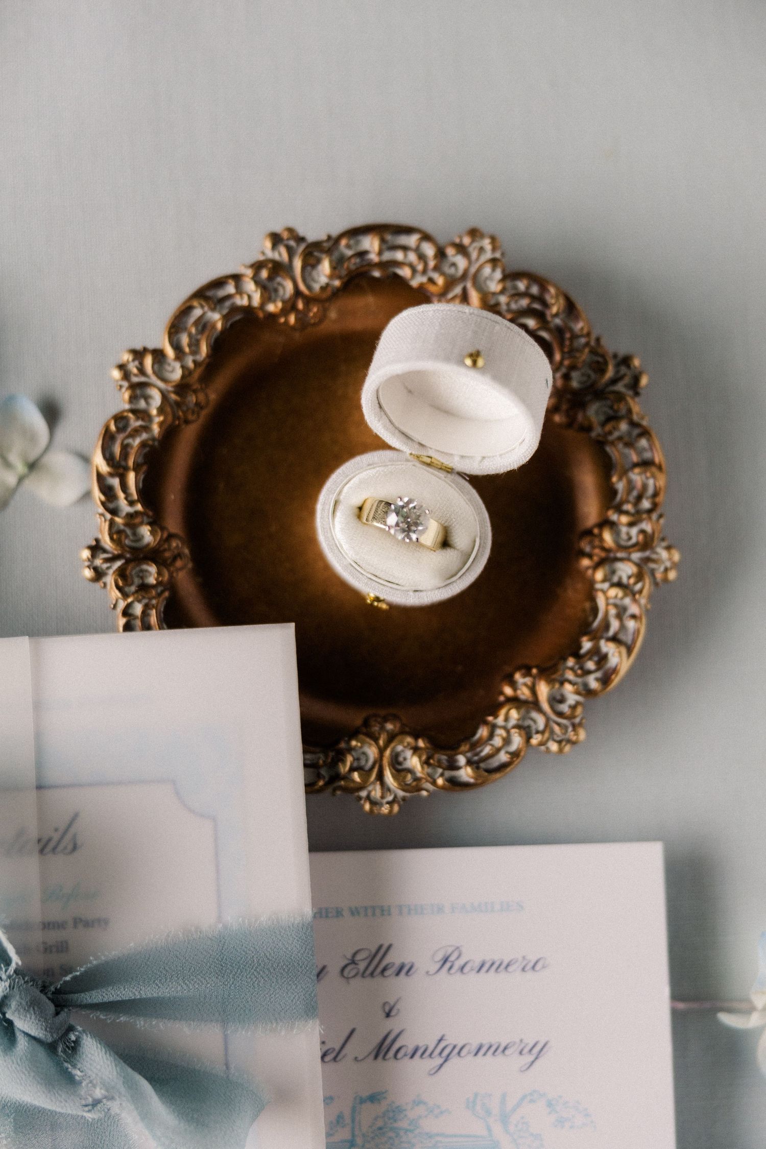 Wedding rings displayed in an ornate white ring box on a decorative gold vintage plate with wedding invitations.