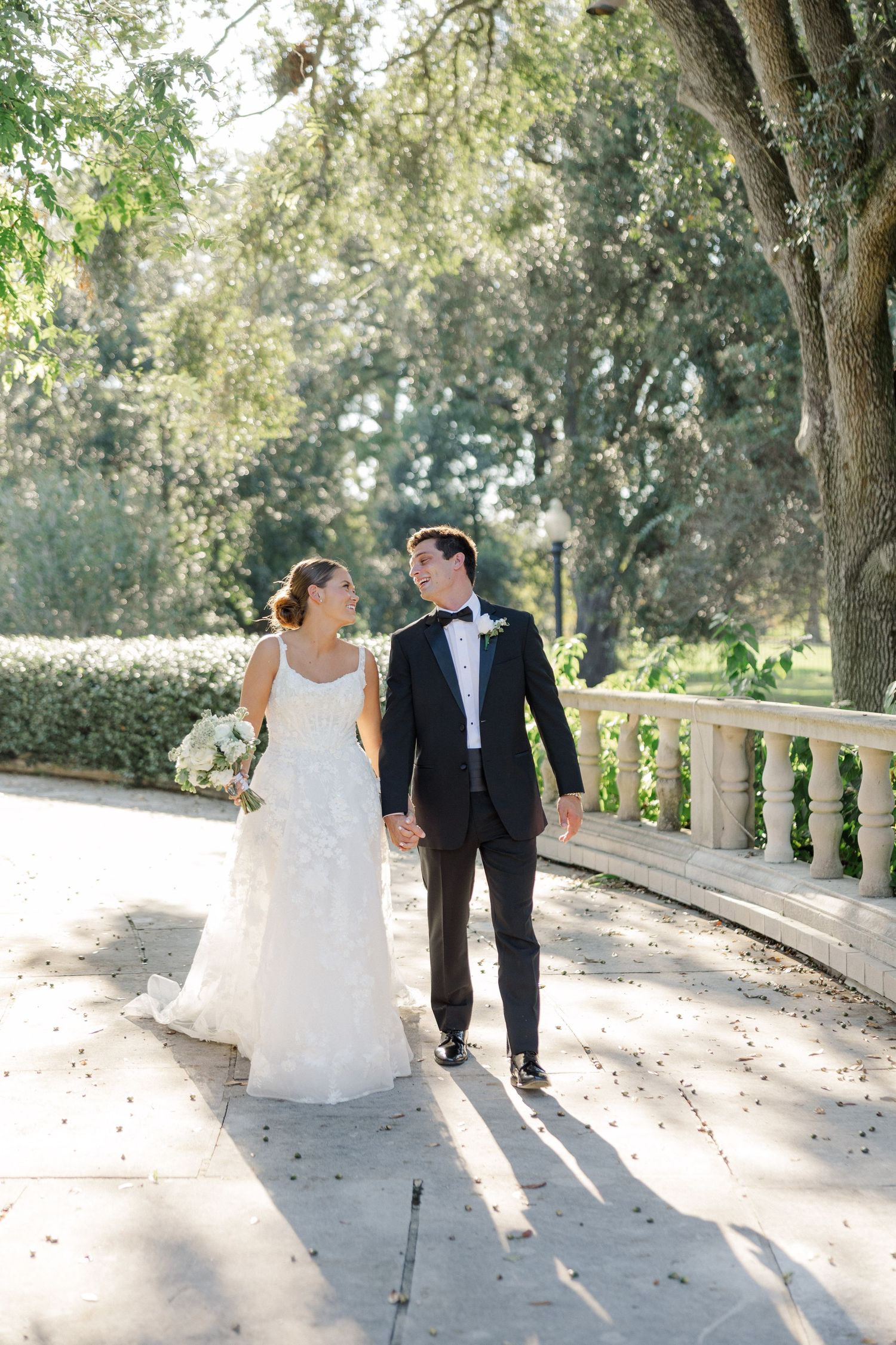 Newlyweds walking and laughing together on a sun-dappled stone pathway.