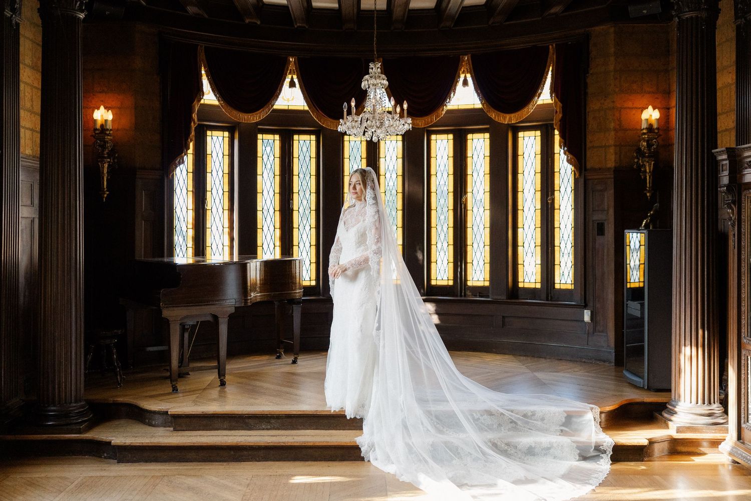 Bride in flowing cathedral veil standing in gothic-style room with stained glass windows.
