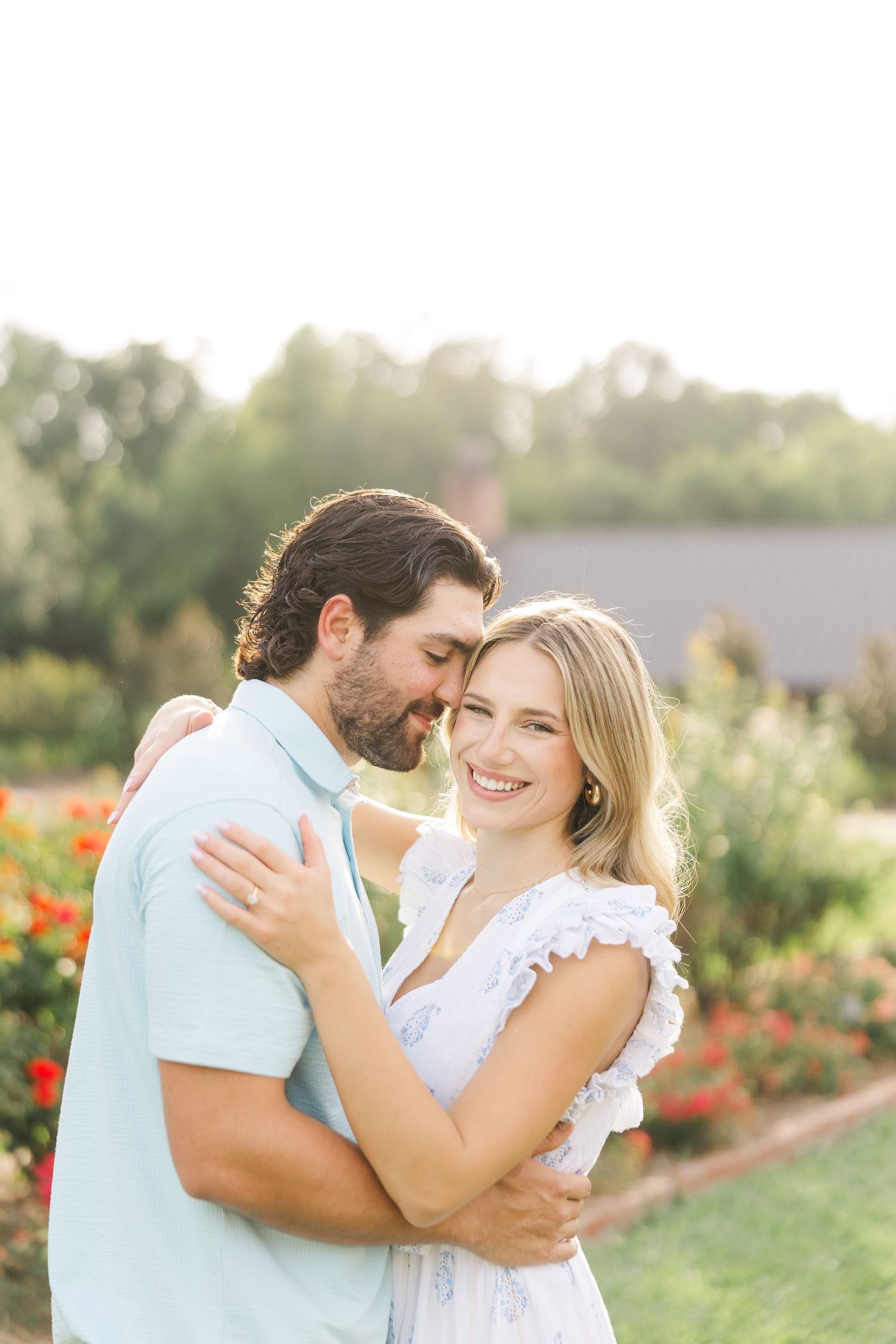 A couple laughs together in a sunlit garden with colorful flowers and greenery surrounding them.