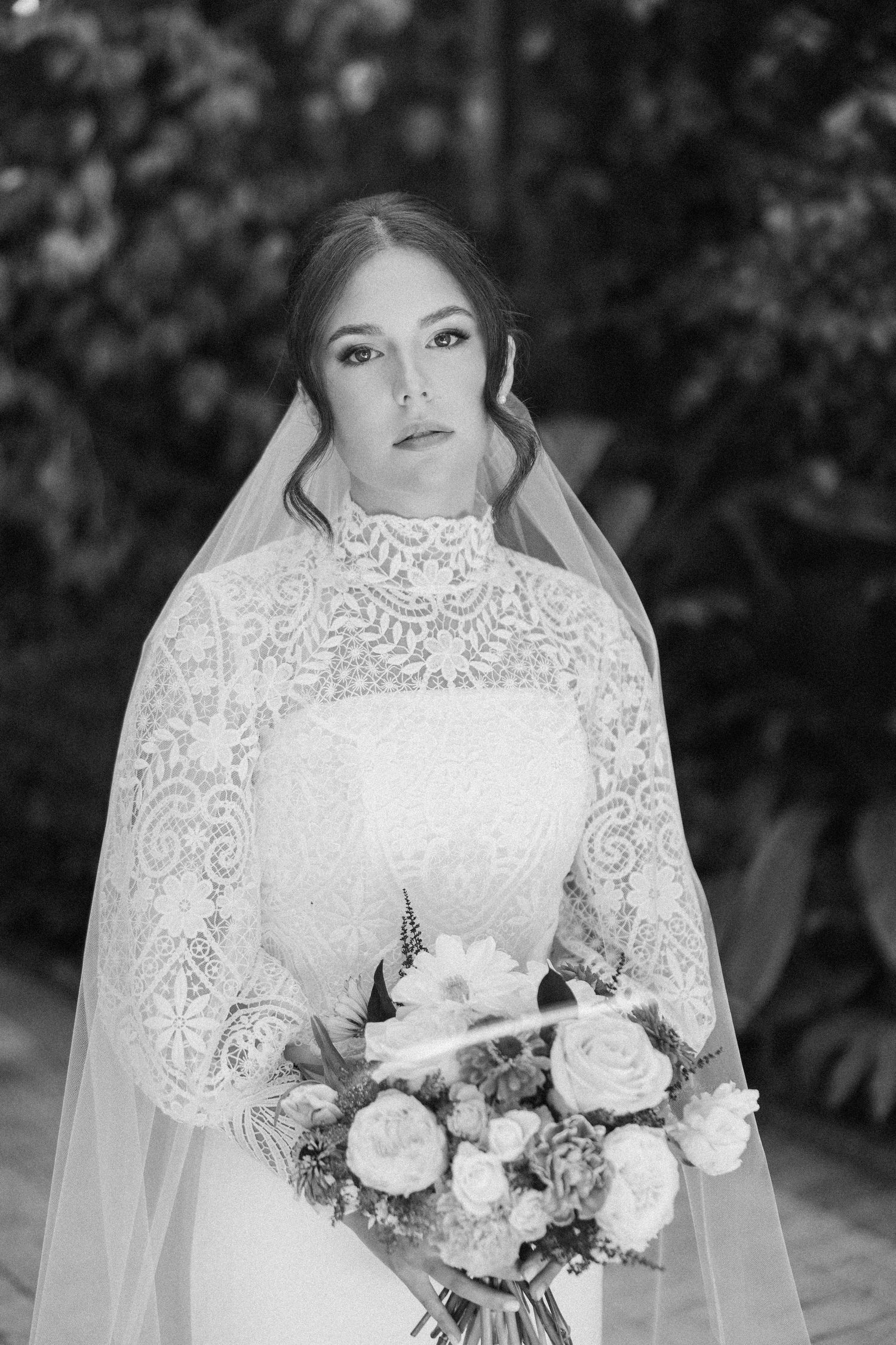 Black and white portrait of bride wearing delicate lace gown and veil while holding floral bouquet.