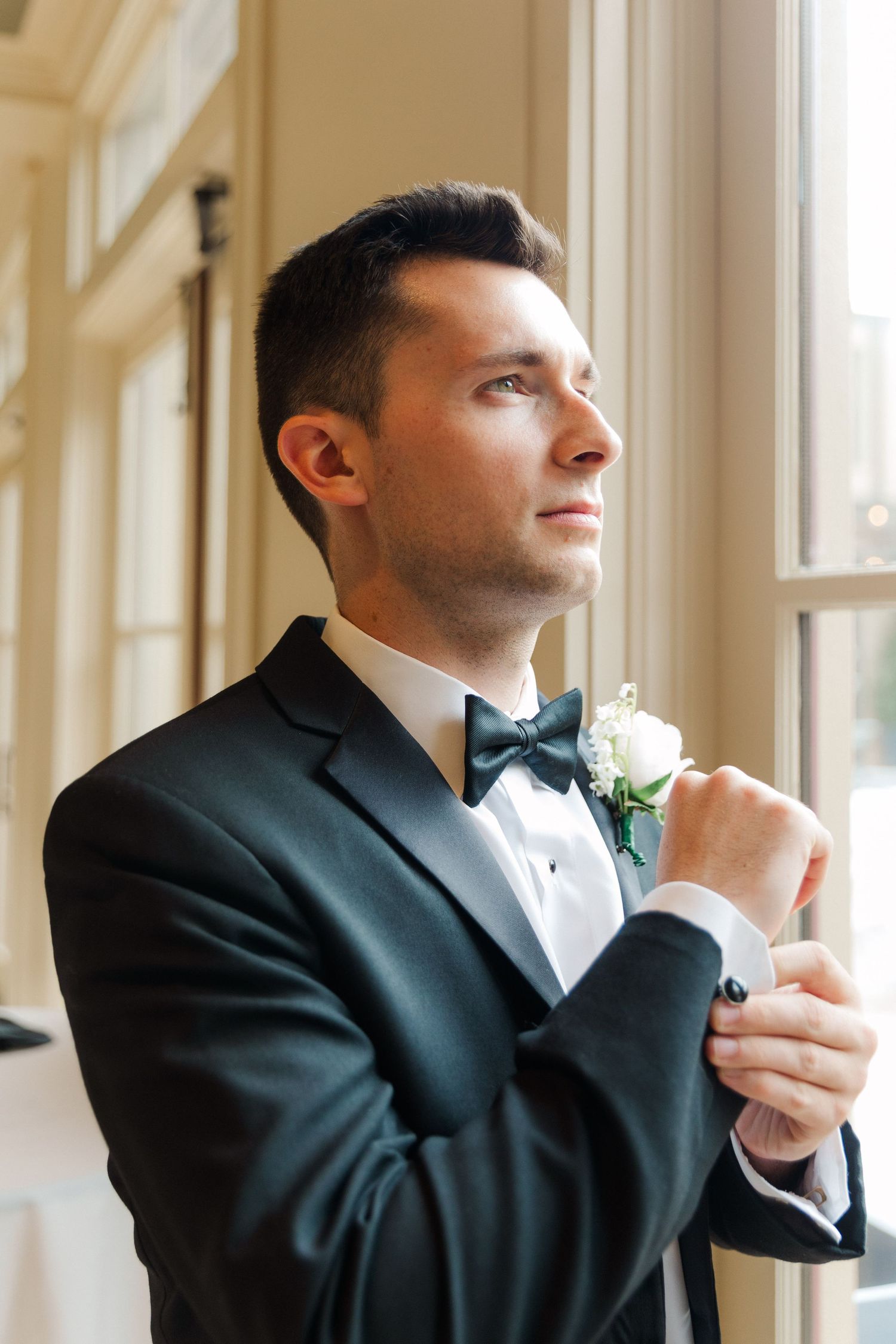 A groom in a black tuxedo adjusts his bowtie while looking out a bright window.