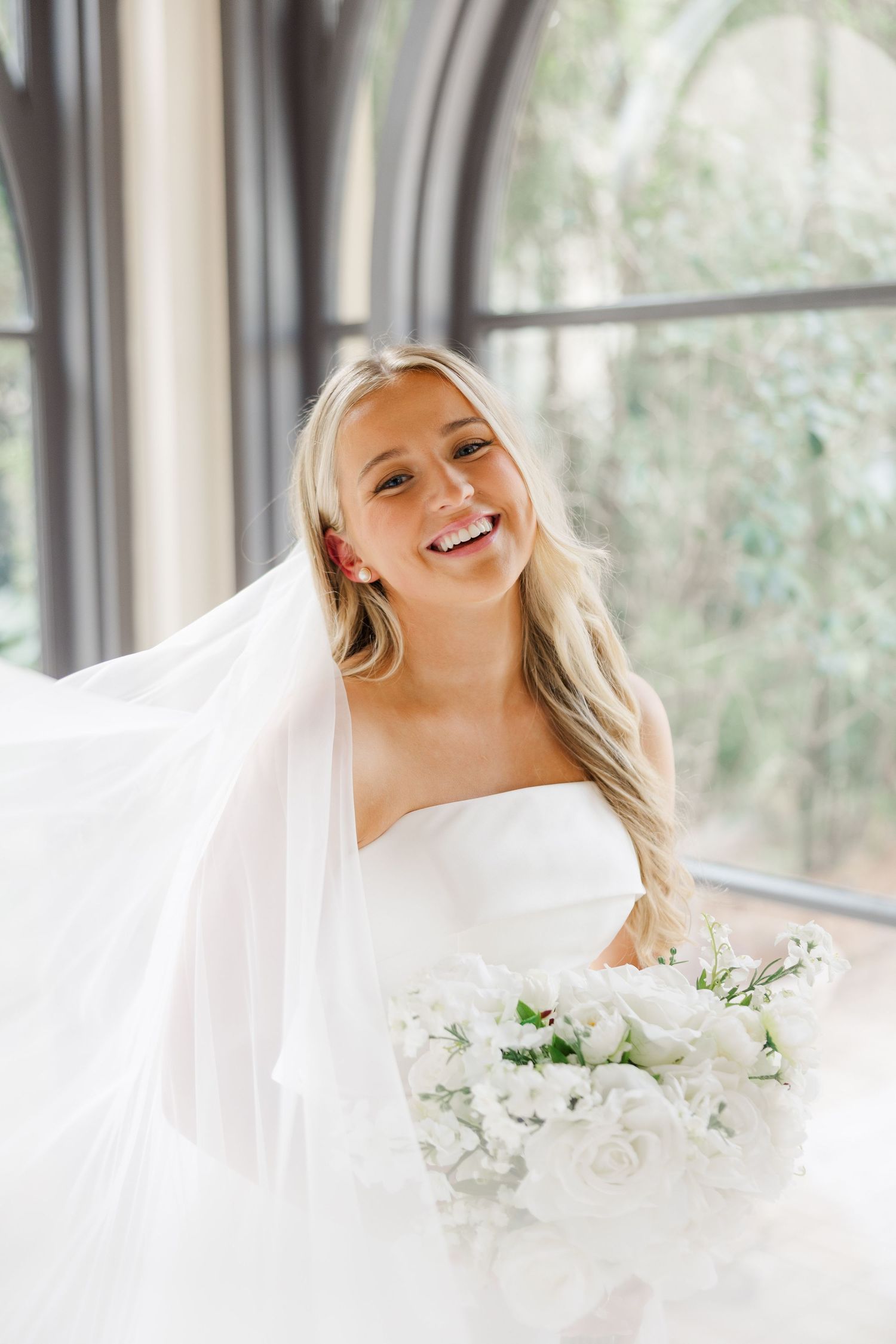 A bride in white dress and veil smiles joyfully while holding a white floral bouquet in a sunlit room.