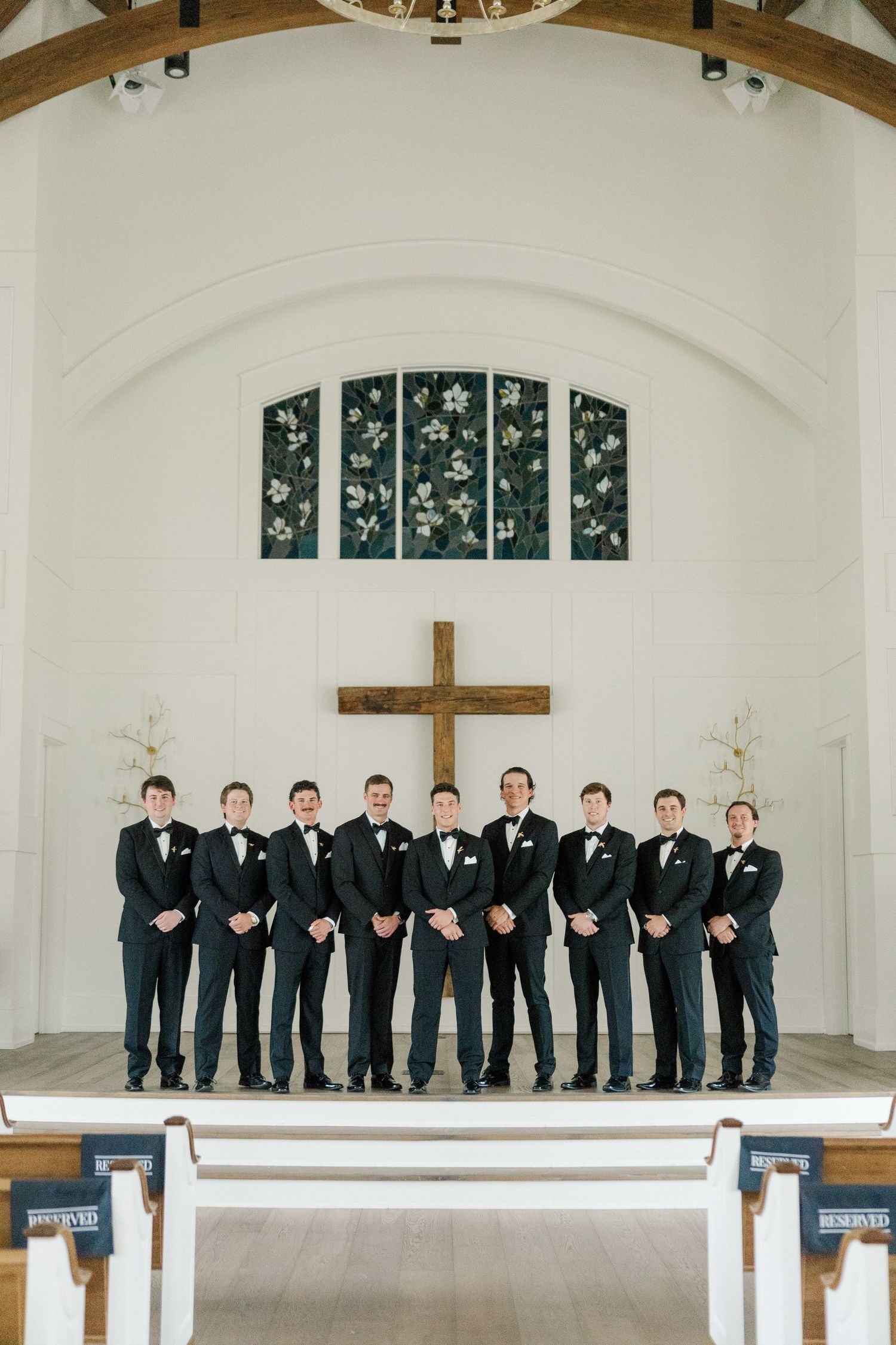 Groomsmen in black tuxedos stand in a line on a church altar with a wooden cross and stained glass windows.