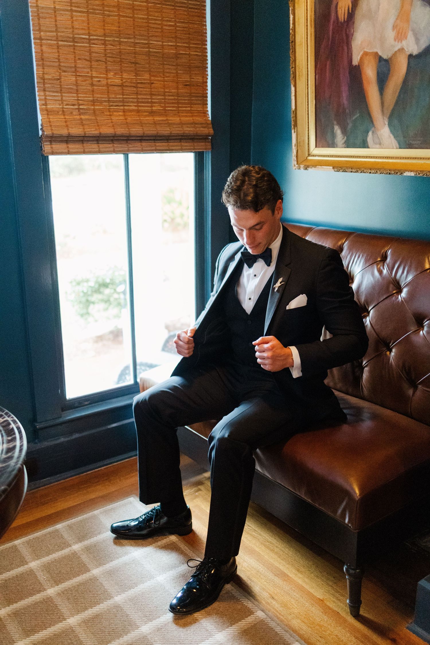A person in a black suit sits thoughtfully on a brown leather couch near a window in a room with wooden floors.